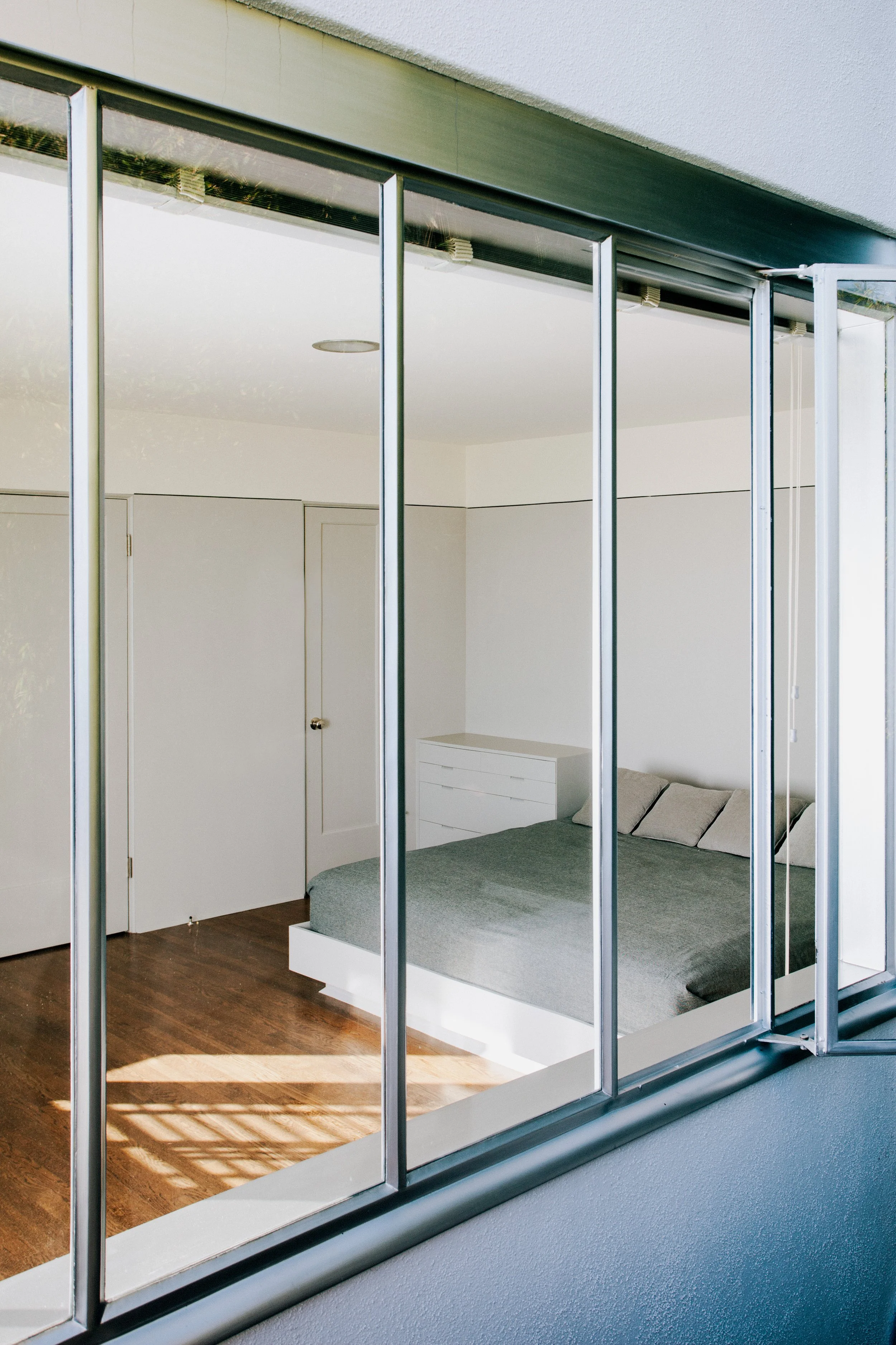 Bright minimalist bedroom viewed through mirrored sliding closet doors, with a white dresser, gray bed, and wood flooring.