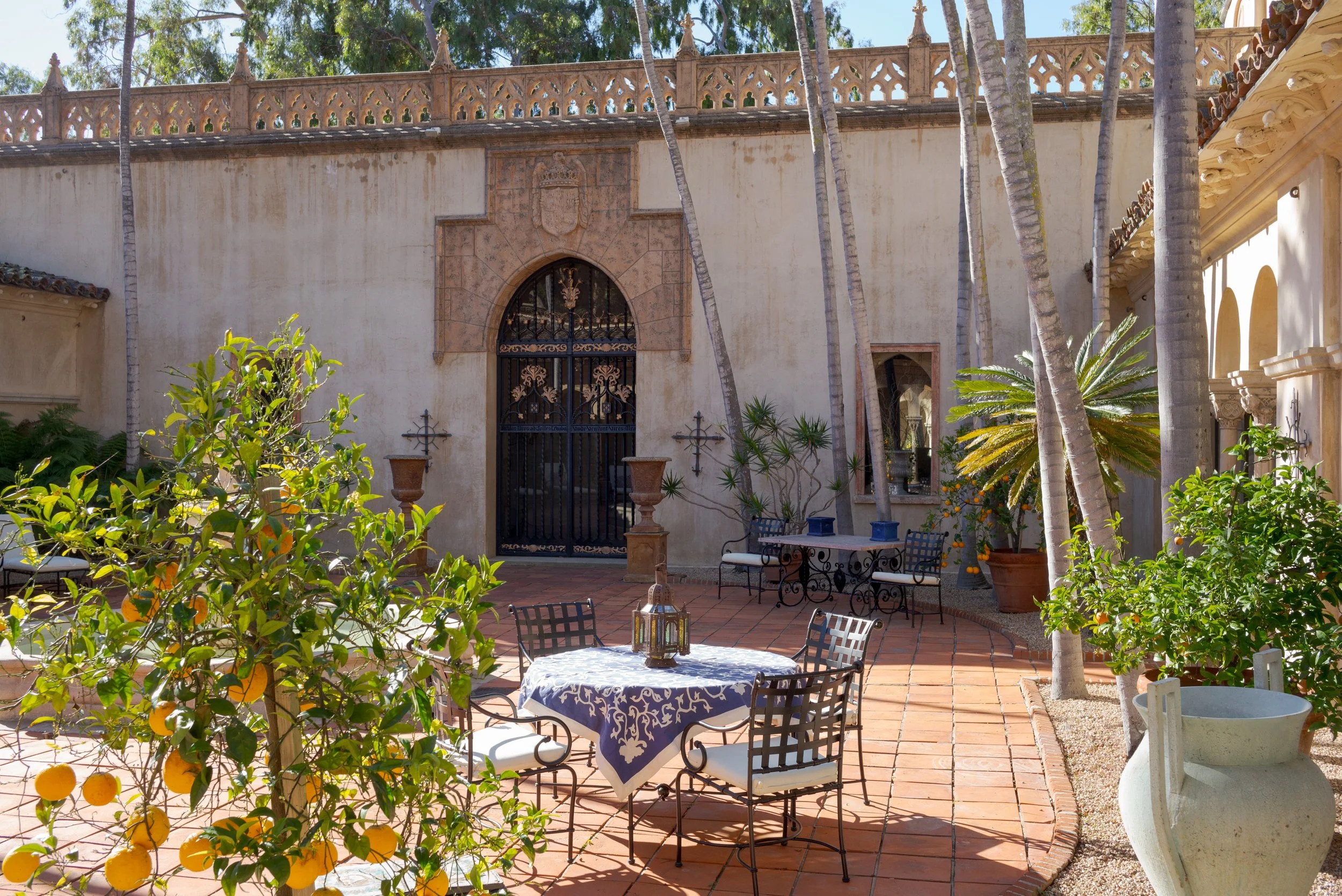 Sunlit Spanish-style courtyard with orange trees, palm trees, ornate stucco walls, arched doorway, and patio dining furniture