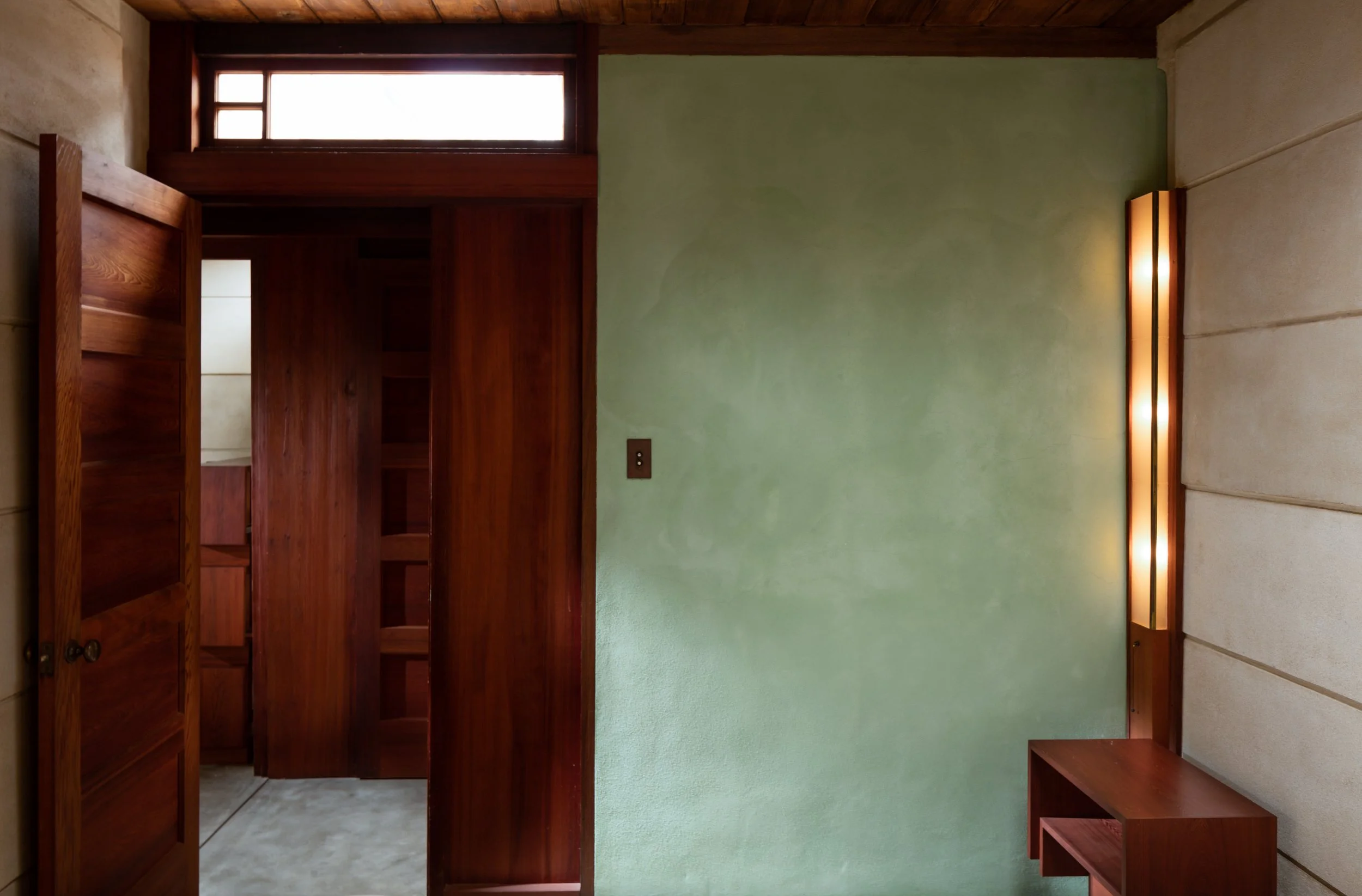 Mid-century modern interior with a green plaster wall, exposed wood ceiling, open wooden doors, built-in shelving, and a vertical wall sconce.