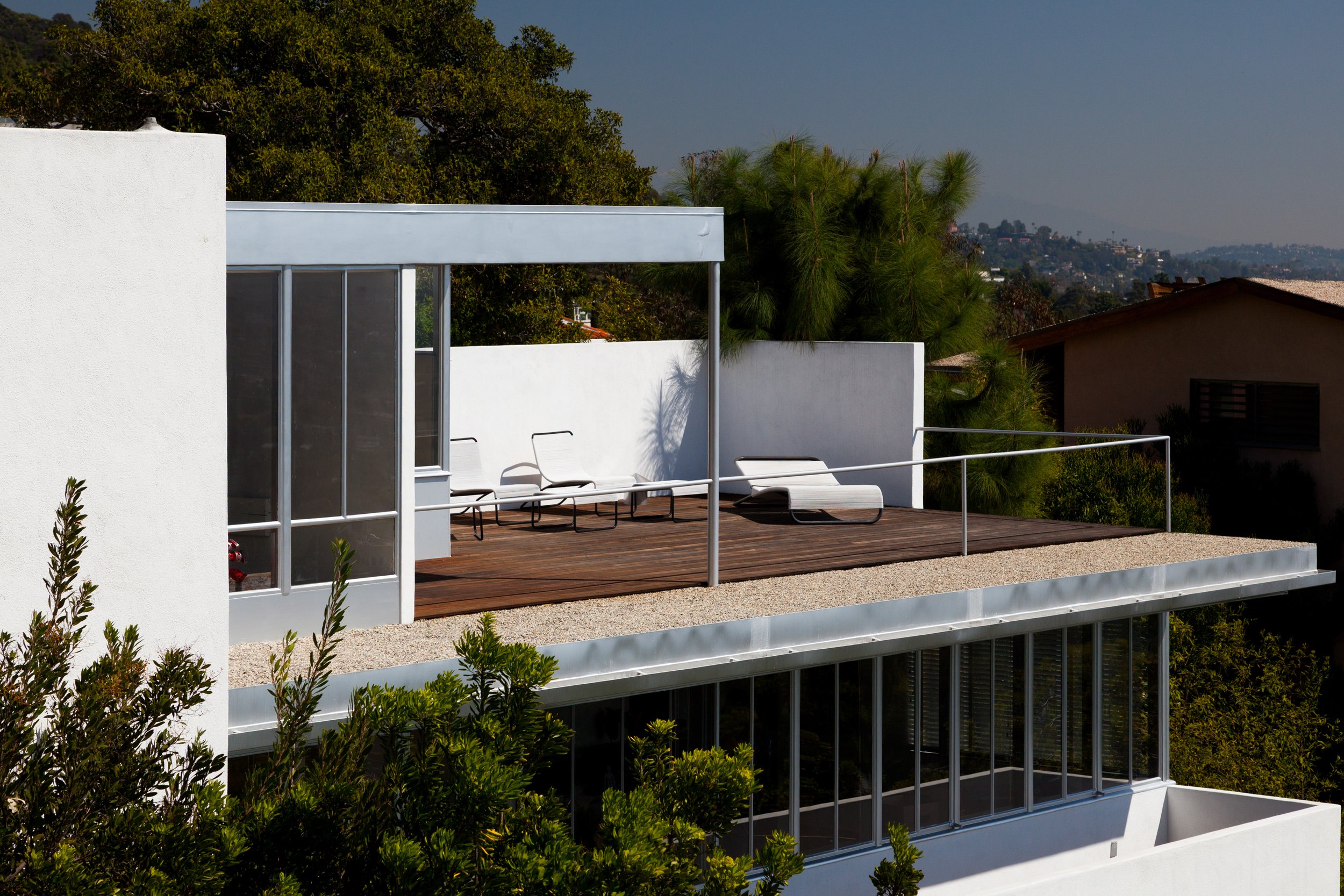 Modern white hillside home with rooftop deck, lounge chairs, trees, and city view
