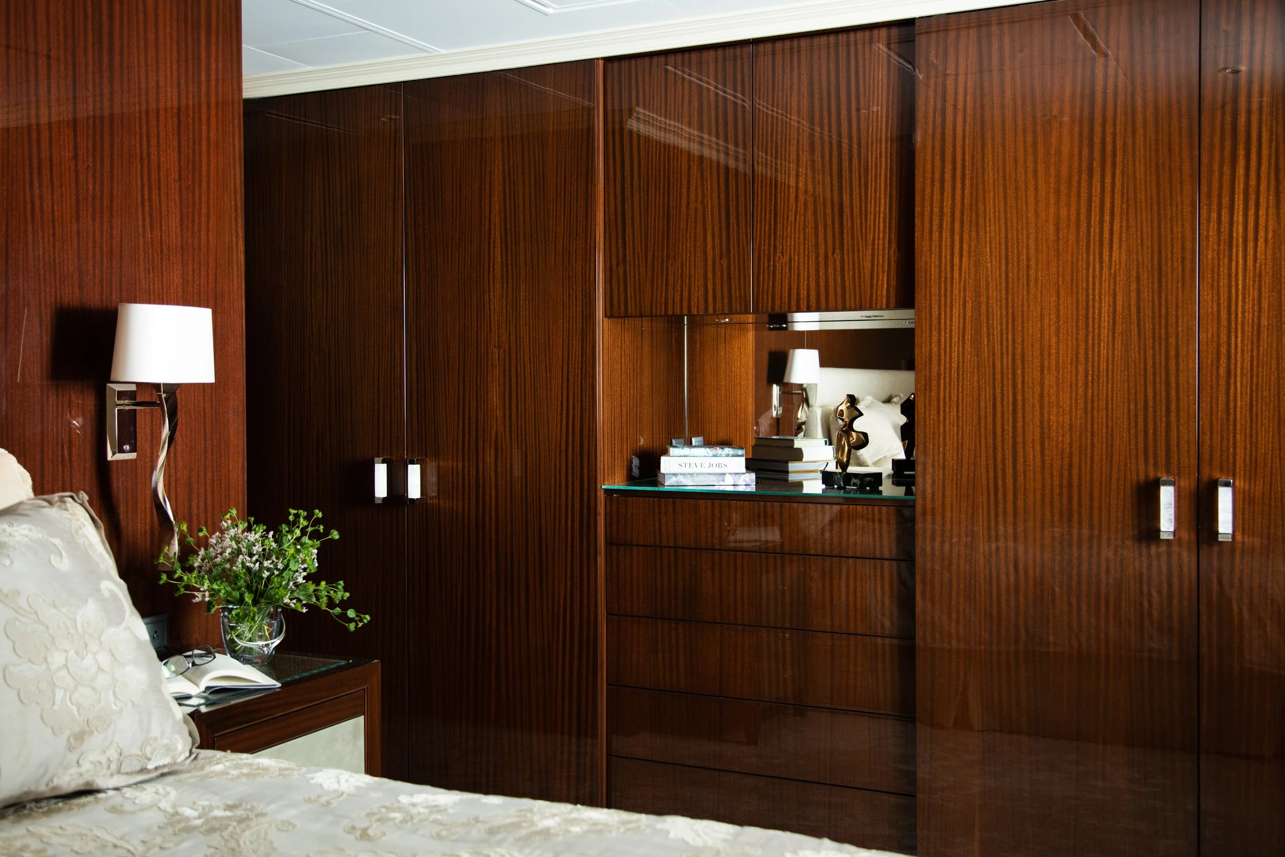 Luxurious bedroom with dark wood paneling, built-in wardrobe, mirrored shelving, bedside lamp, flowers, and a neatly made bed