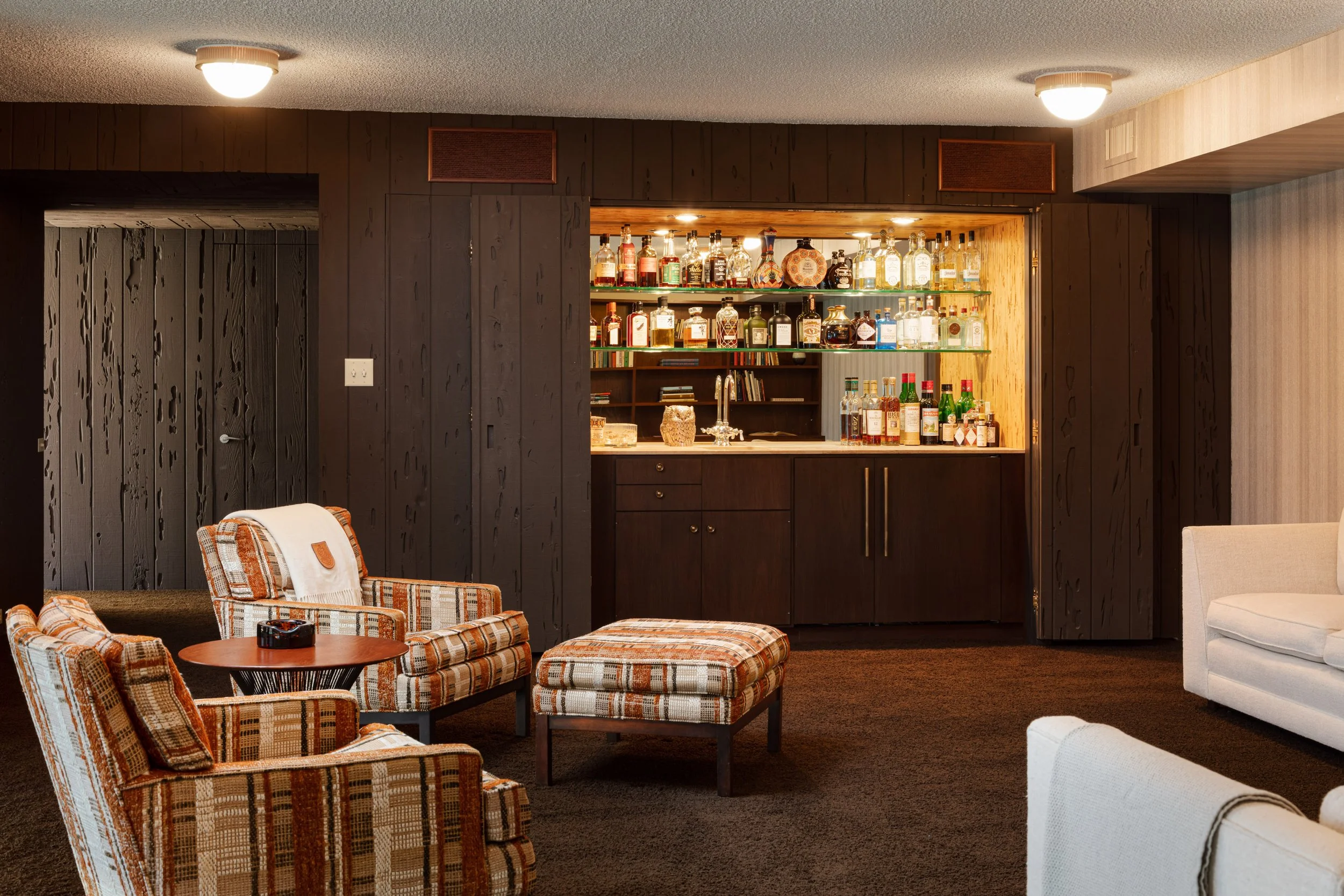 Retro basement lounge with a well-stocked built-in liquor bar, wood-paneled walls, patterned armchairs, and ottomans