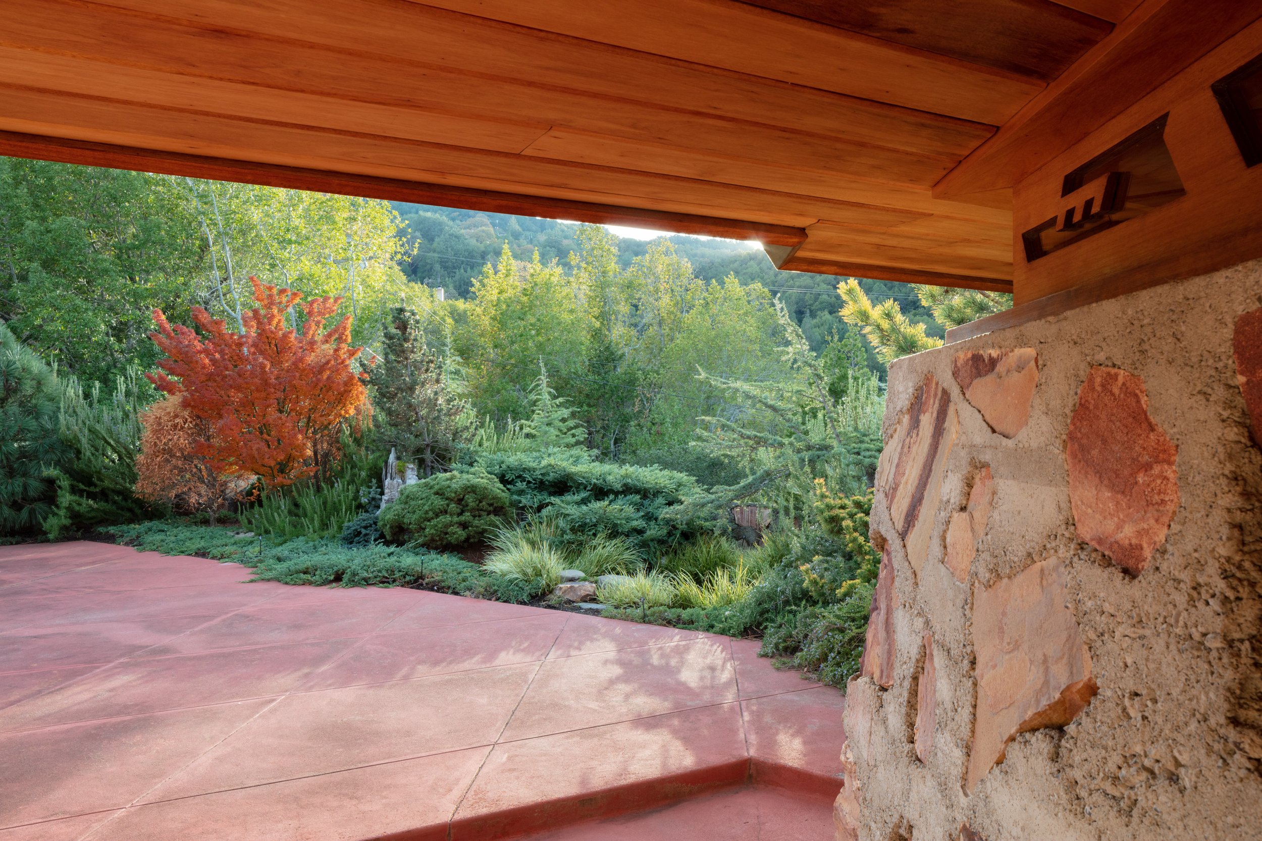 Covered patio with timber roof and stone wall overlooking a landscaped garden and forested hillside
