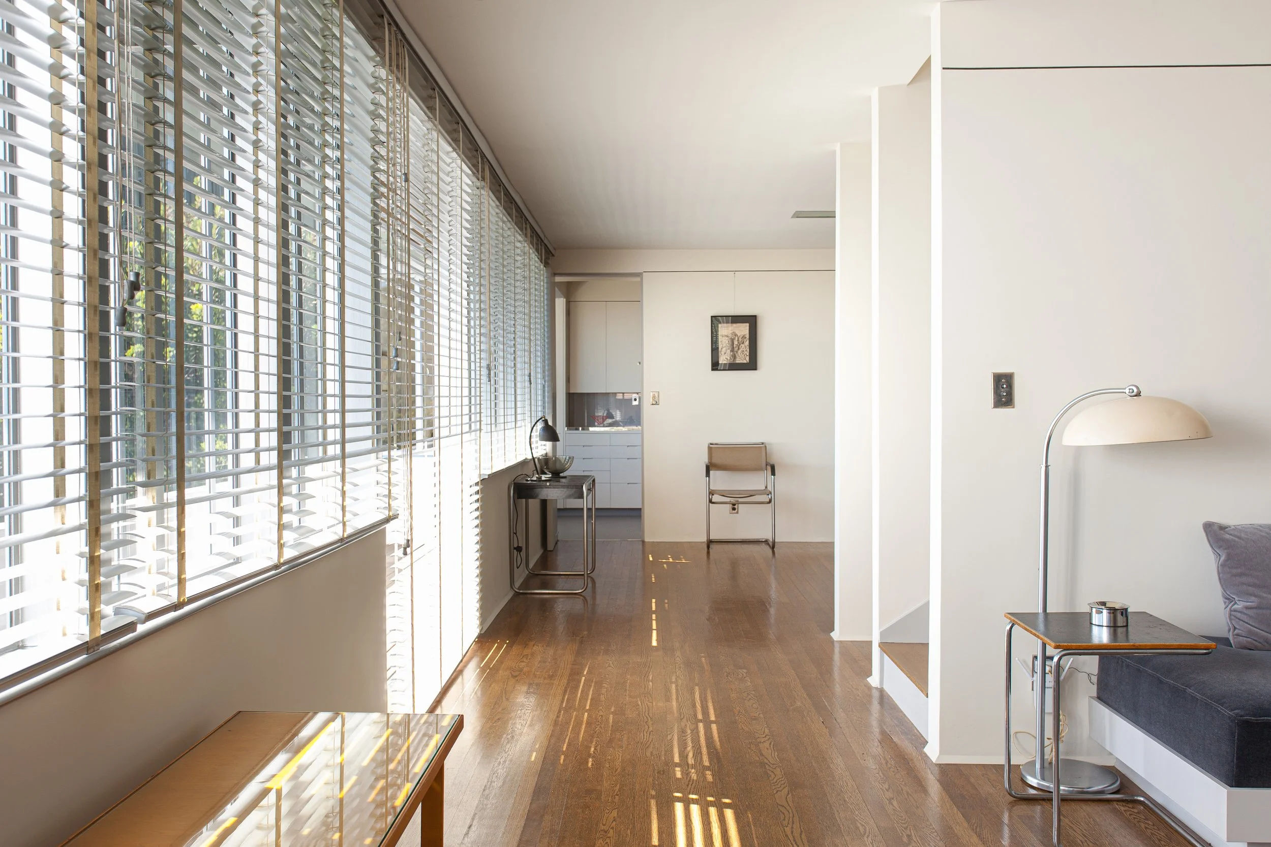 Bright modern living room with wood floors, large windows with venetian blinds, minimalist furniture, and a dome floor lamp