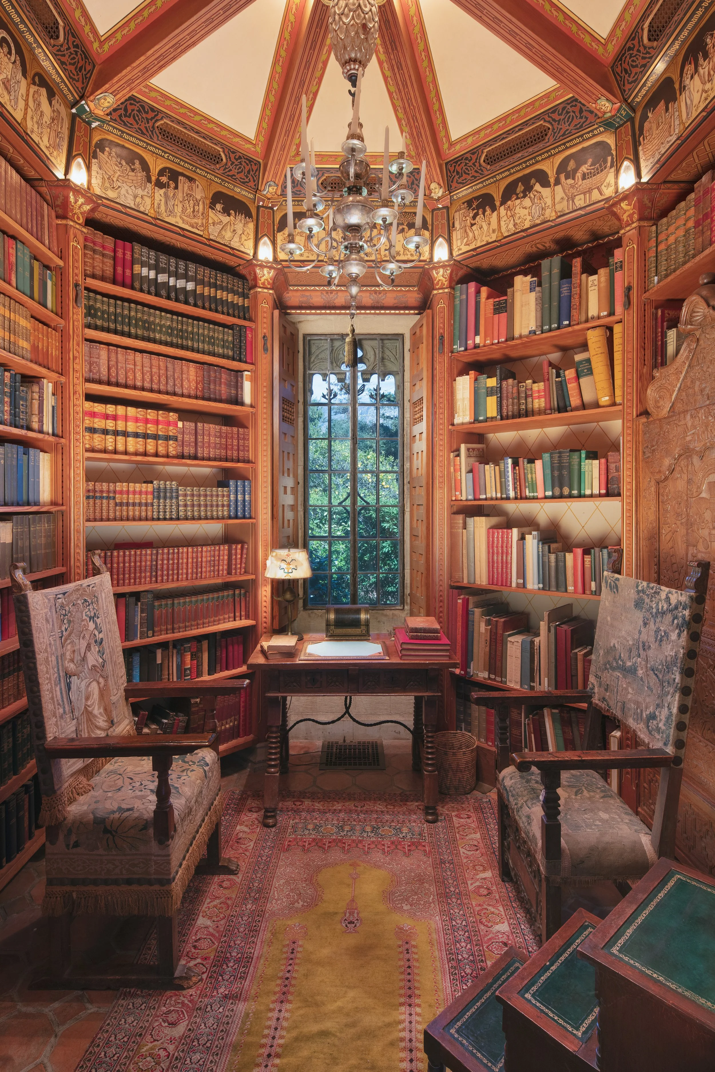 Ornate wood-paneled library filled with antique books, biblical murals, a central chandelier, upholstered chairs, and a patterned Persian rug