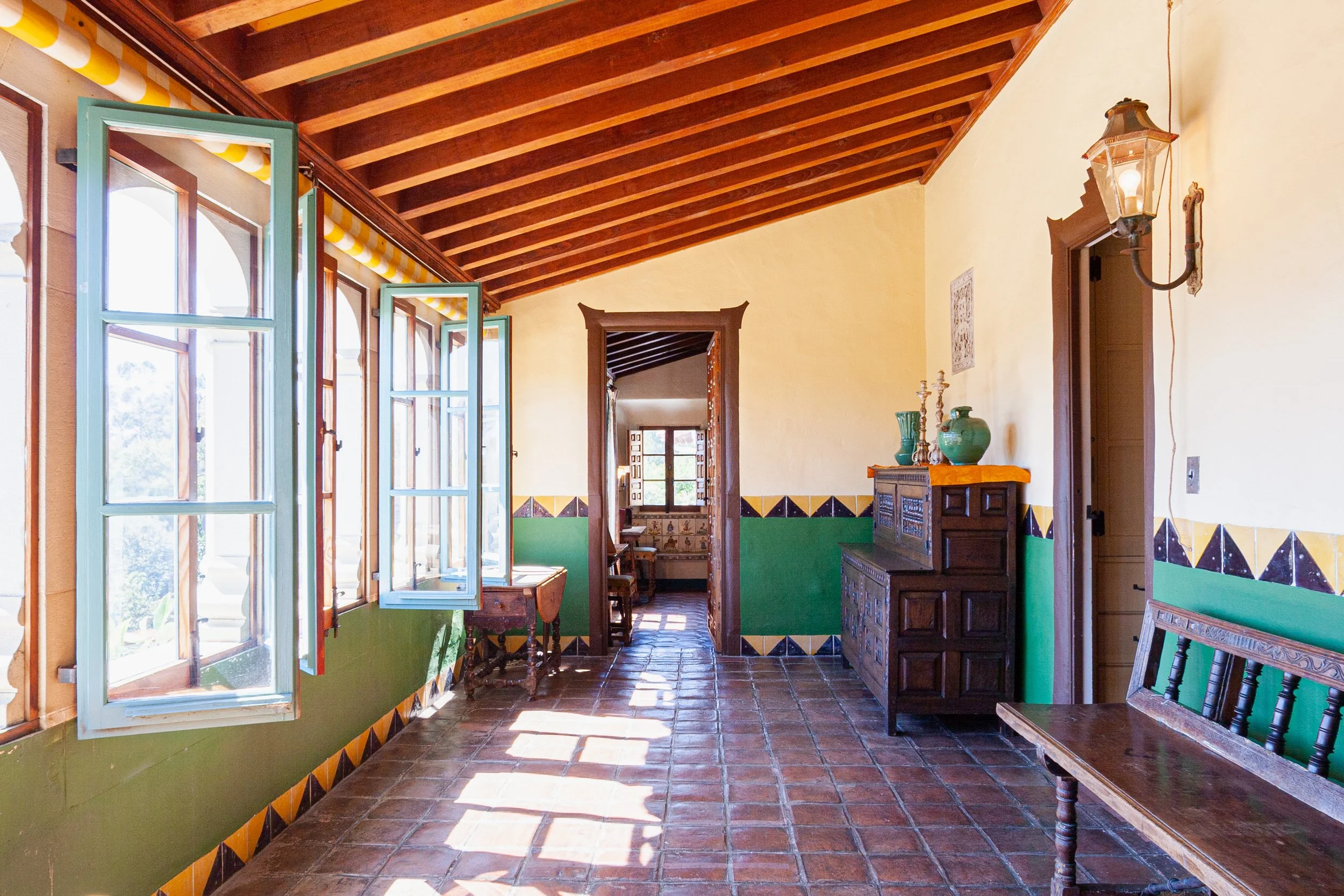 Historic Spanish-style hallway with open turquoise windows, exposed wood-beam ceiling, terracotta tile floor, decorative ceramic tile walls, antique cabinet, and wall lantern