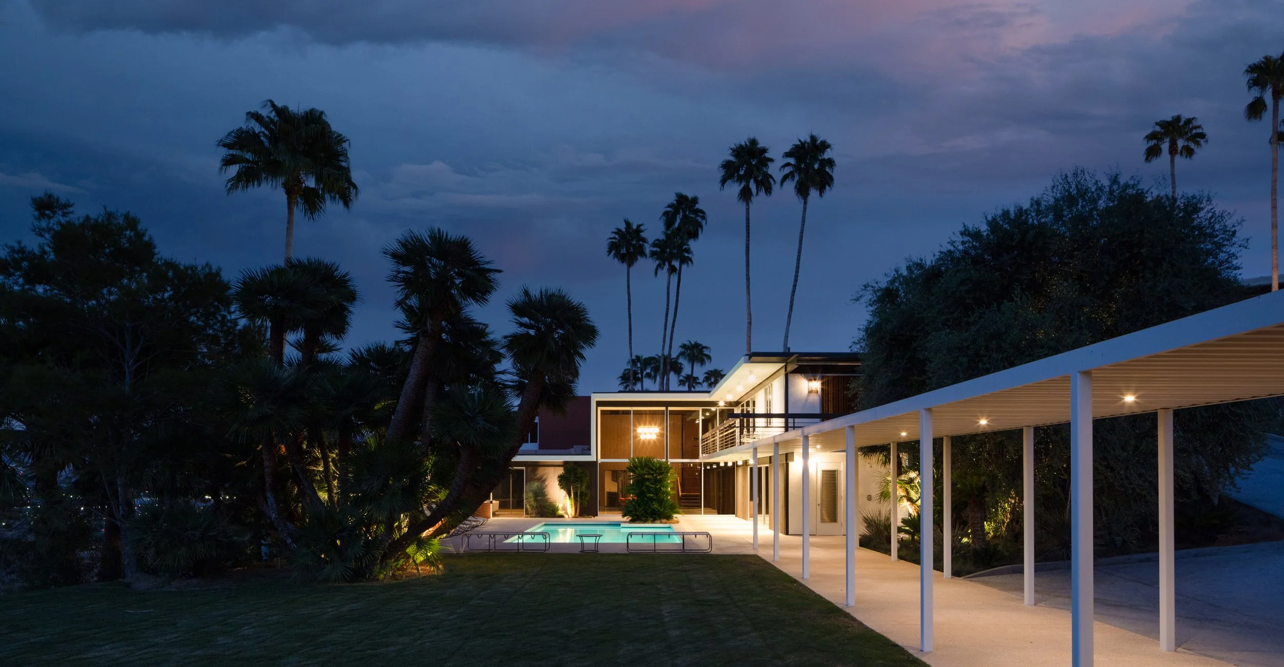 Modern midcentury house with a lit swimming pool, covered patio, and palm trees at dusk