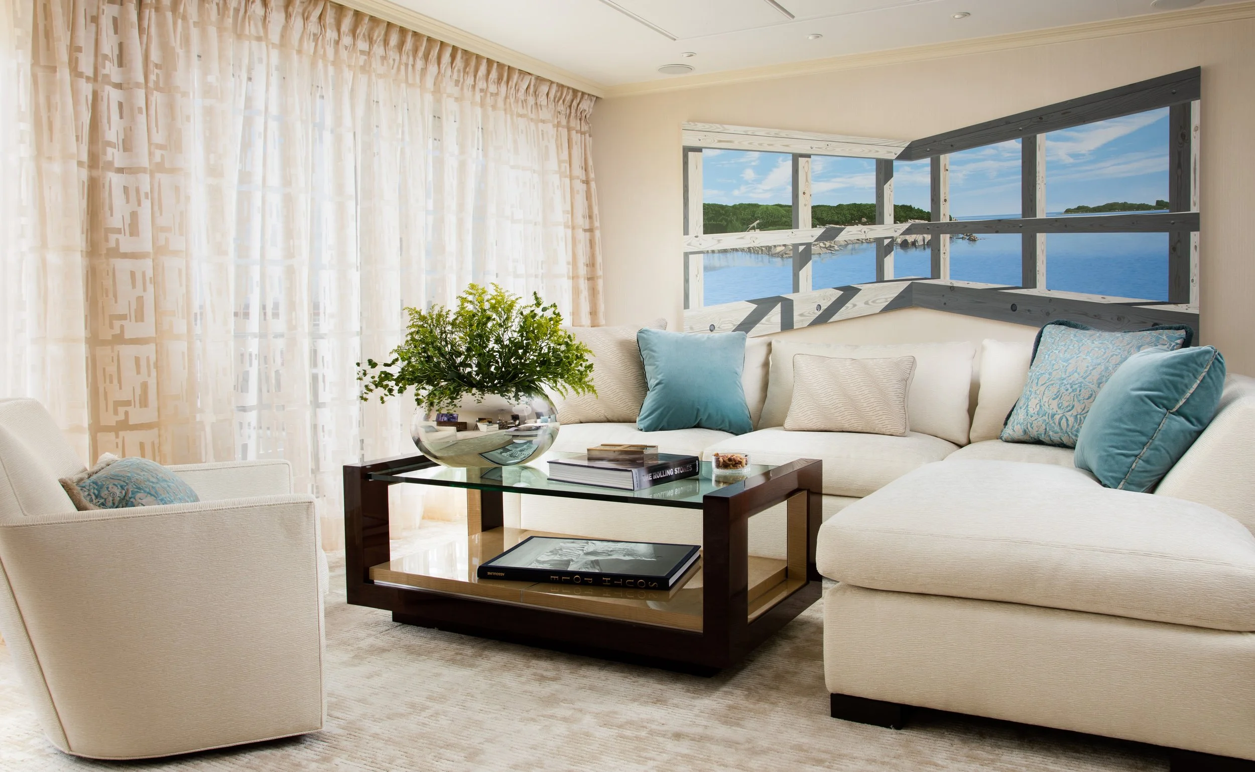 Bright coastal living room with cream sofas, blue accent pillows, glass coffee table, potted plant, and ocean views through large windows