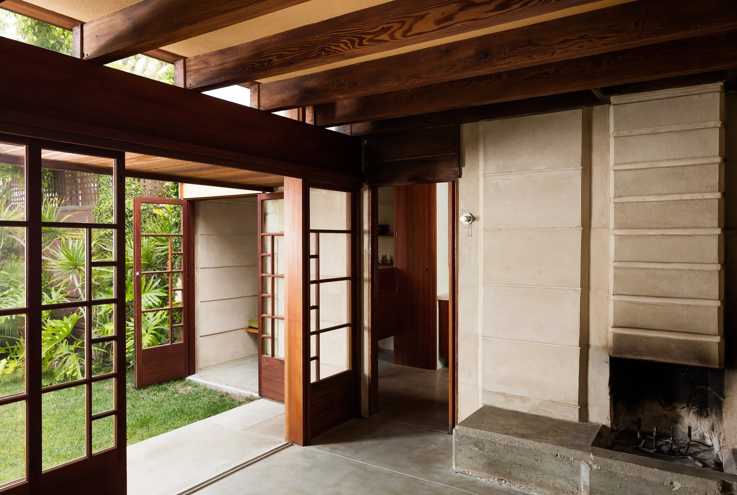 Midcentury modern interior with exposed wood ceiling beams, glass doors opening to a lush garden, and a large concrete fireplace.