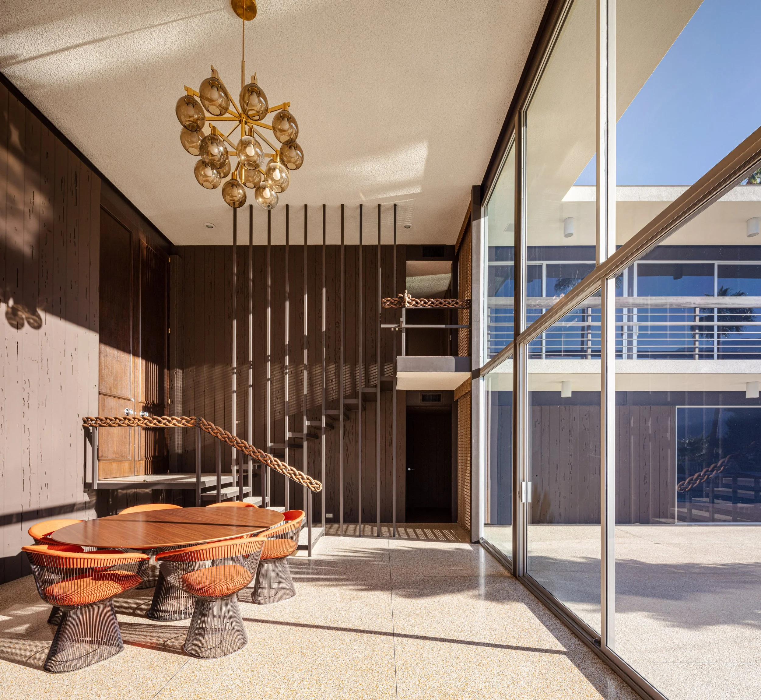 Midcentury modern dining room with orange wire chairs, wood table, sculptural chandelier, spiral staircase, and floor-to-ceiling windows