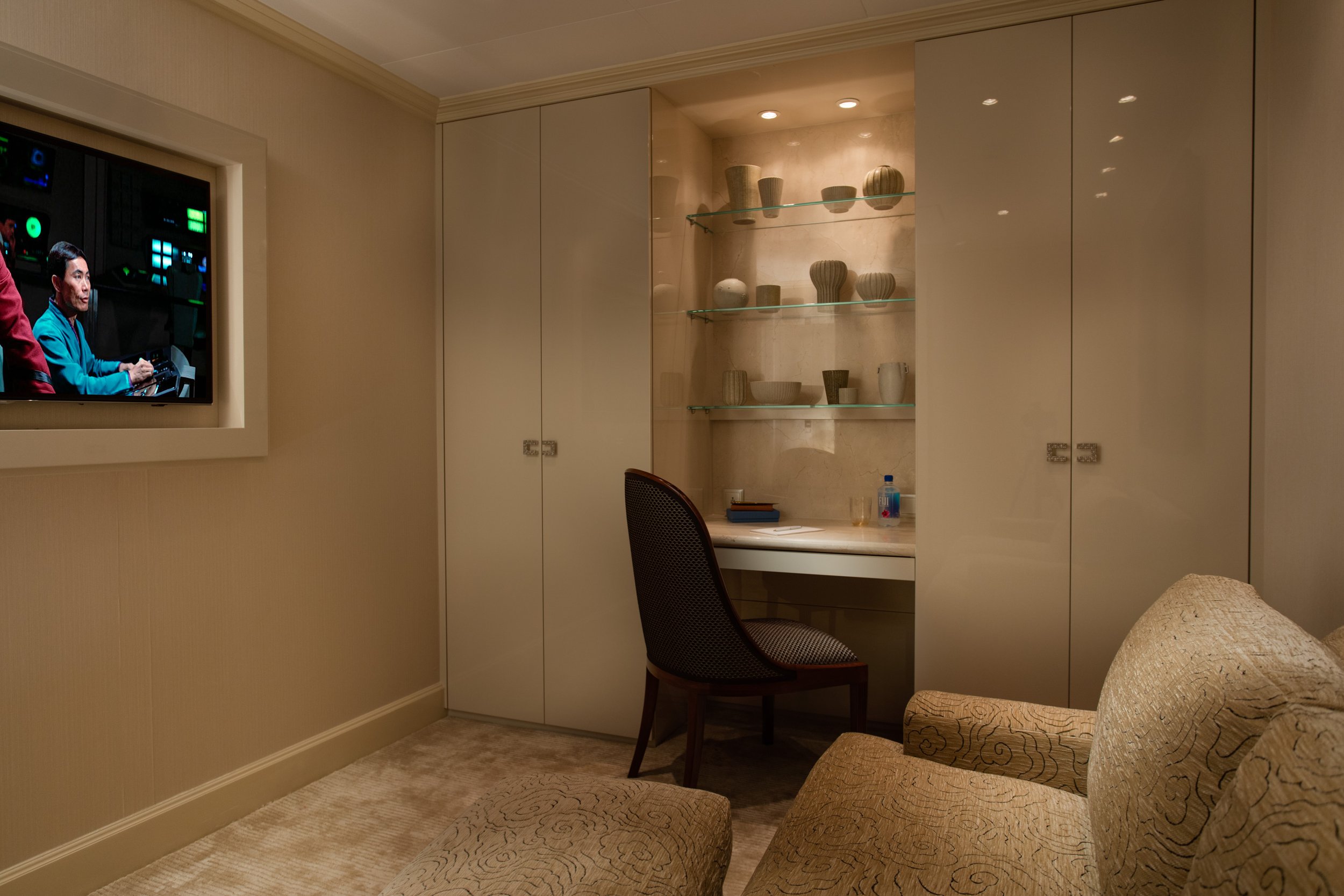 Warmly lit luxury hotel room with a desk, chair, decorative pottery shelves, built-in cabinets, television, and patterned armchairs.