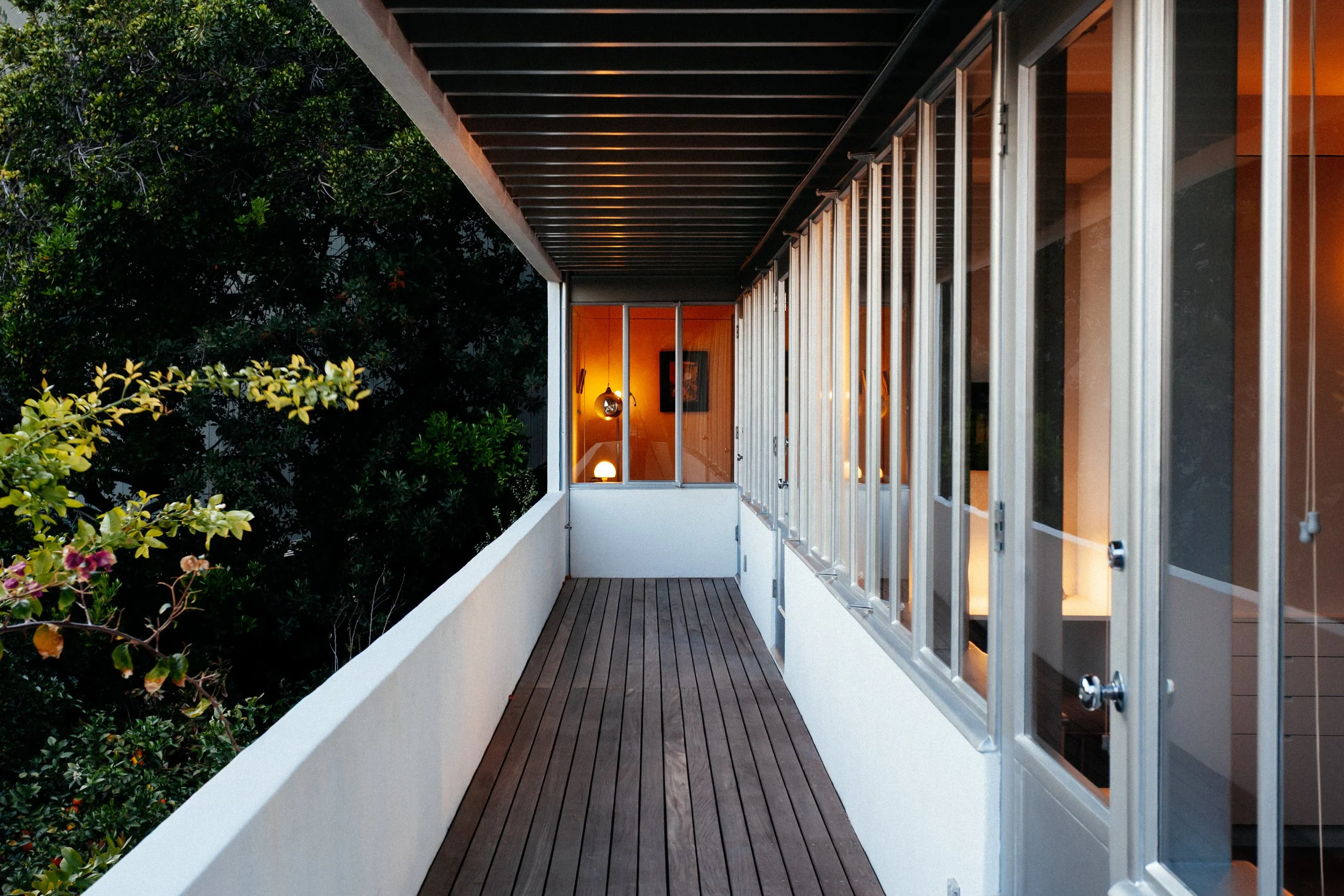 Modern home balcony with wood decking, glass doors, warm interior lighting, and lush greenery