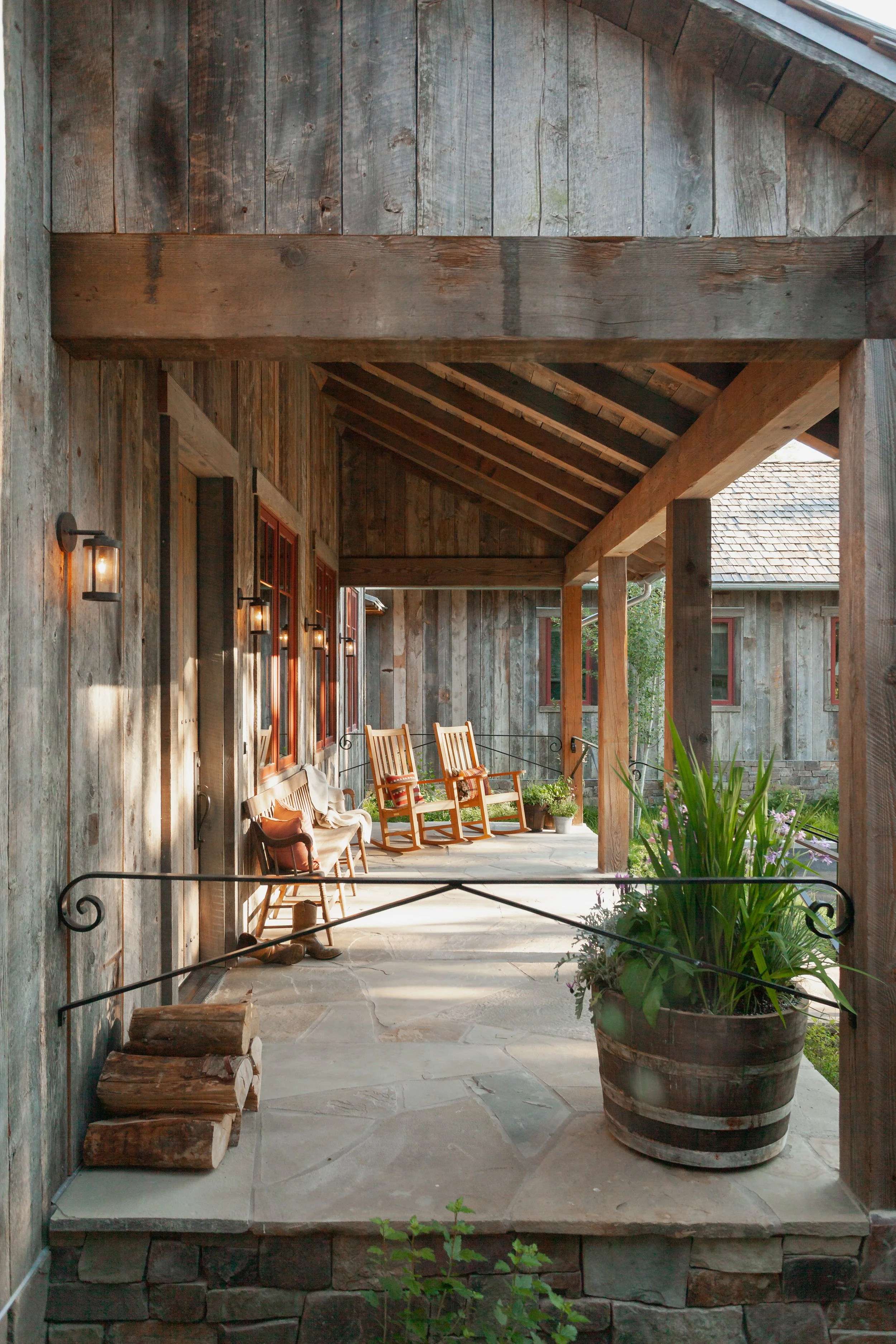 Rustic covered porch with weathered wood walls, exposed beams, rocking chairs, lanterns, potted plants, and a stone floor.