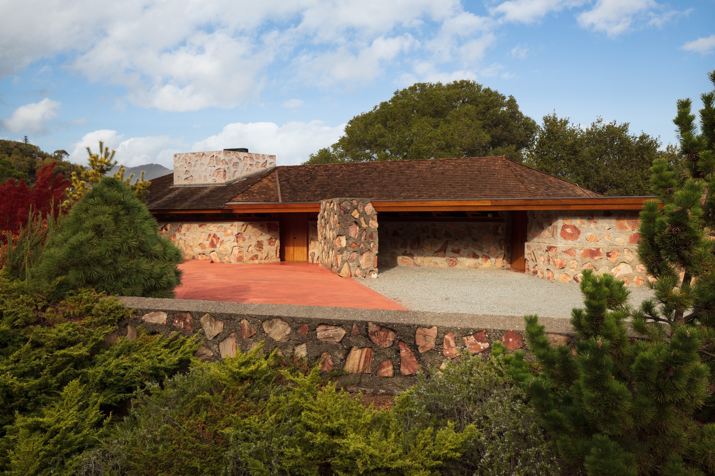 Low stone-and-wood building with a tiled roof, surrounded by pine trees and shrubs beneath a blue sky.