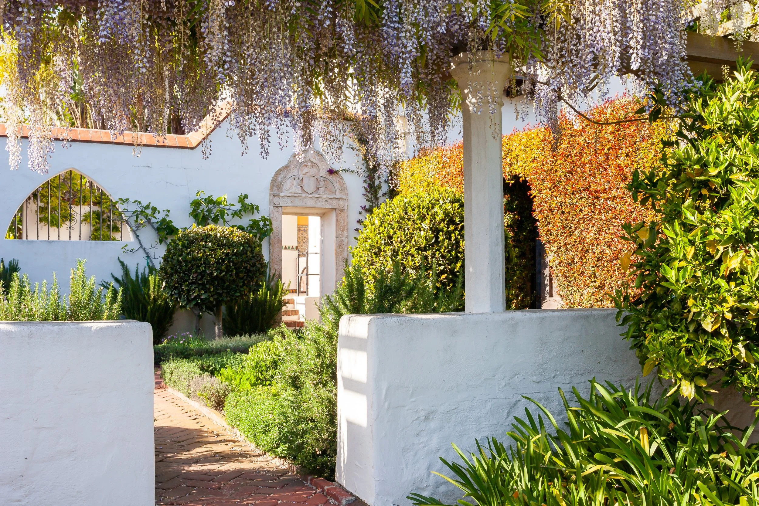 Lush courtyard garden with cascading purple wisteria, white stucco walls, brick pathway, and ornate doorway