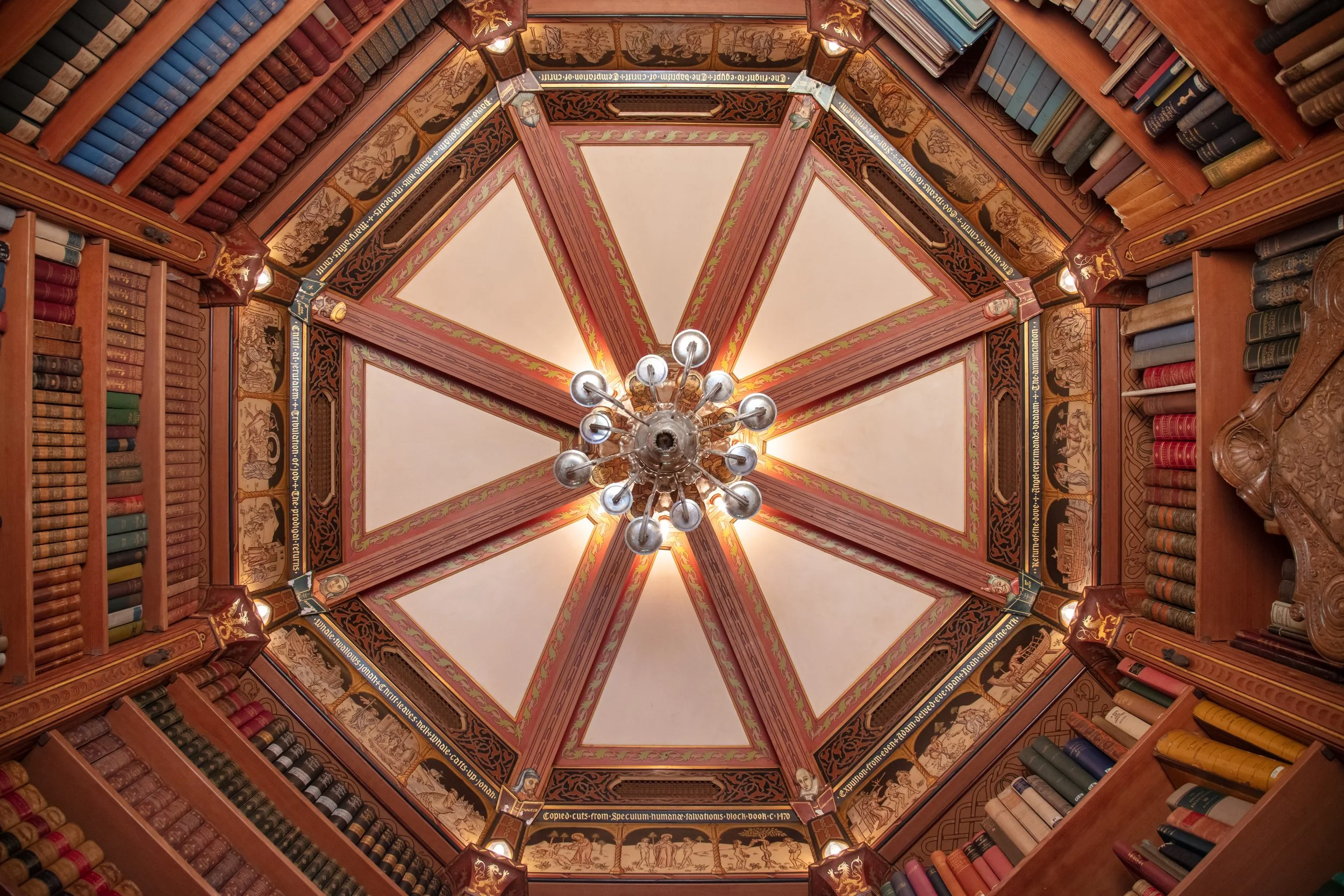 Ornate library ceiling with radial decorative panels, chandelier, carved woodwork, and bookshelves filled with antique books