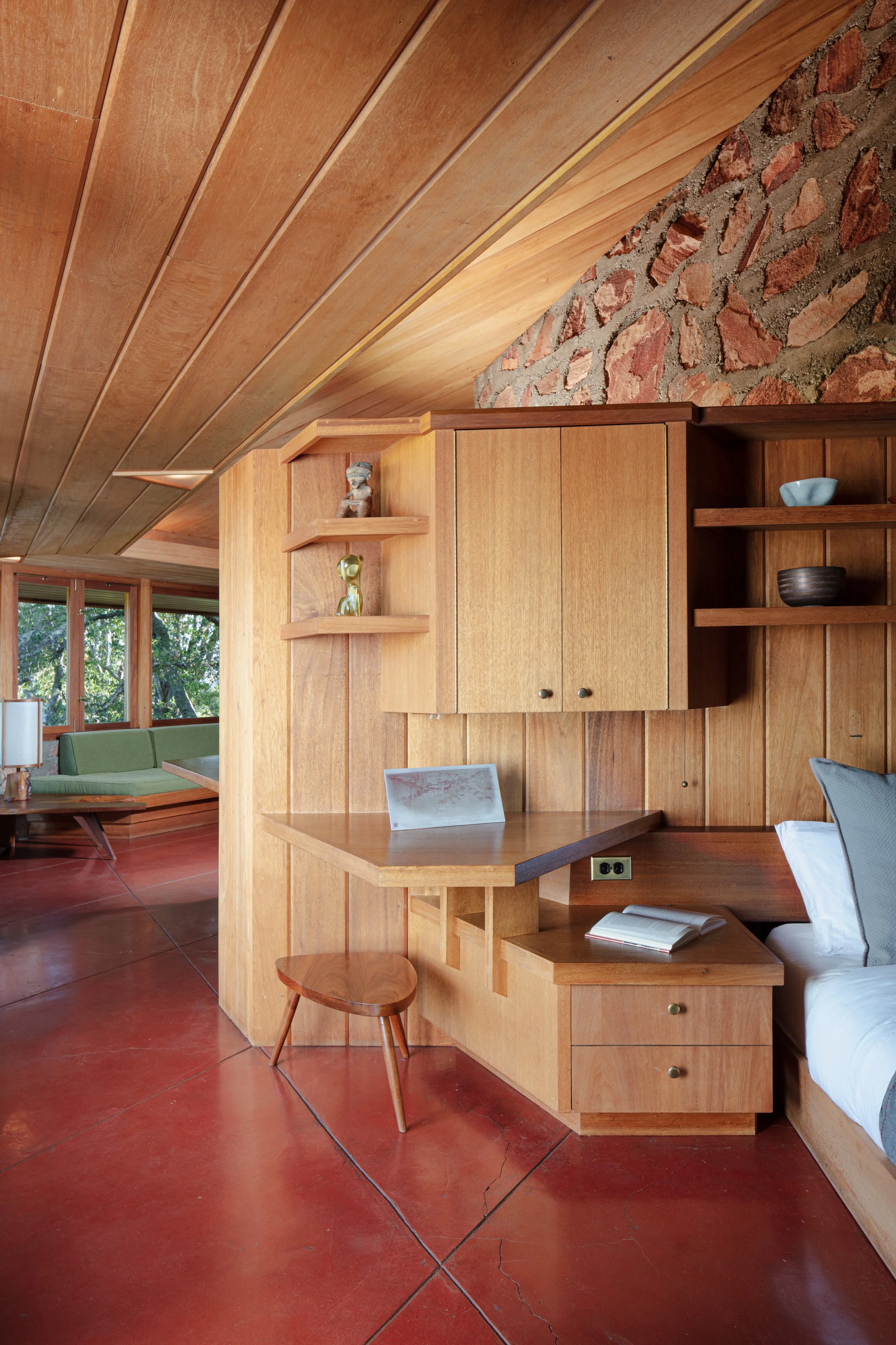 Midcentury modern wood-paneled bedroom with built-in cabinets, open shelving, desk, bed, red tile floor, and exposed brick wall