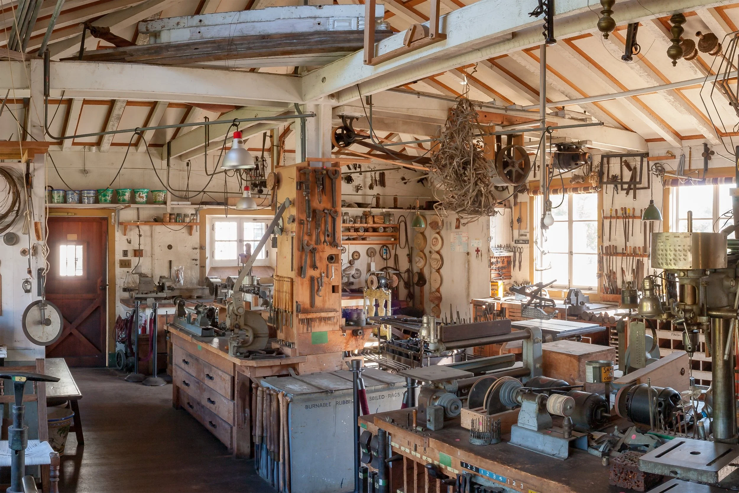 Interior of a historic metalworking workshop filled with lathes, drill presses, hand tools, machinery, and hanging work lights