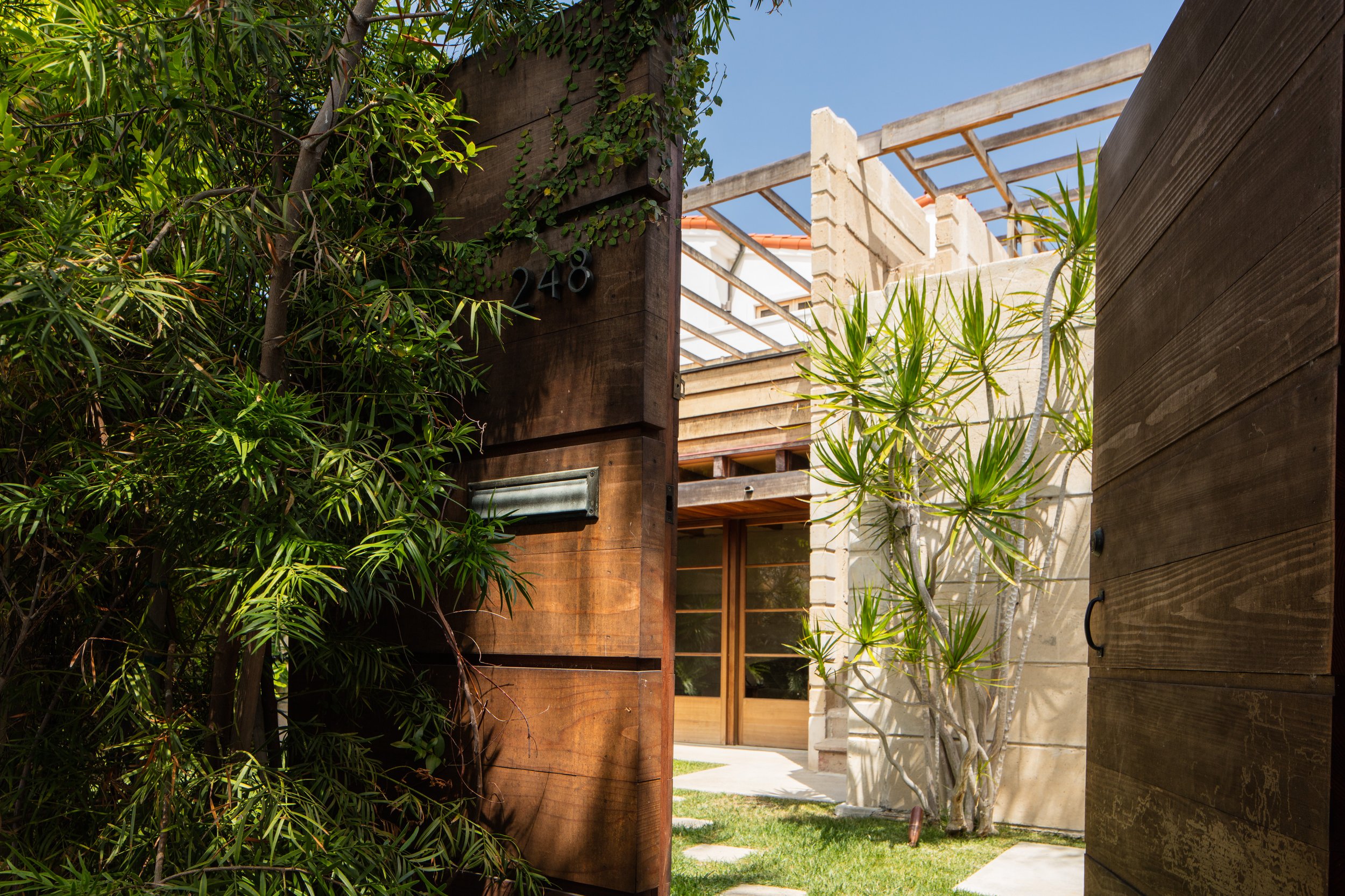 Wooden gate marked 248 opening to a lush courtyard with tropical plants and a modern concrete home