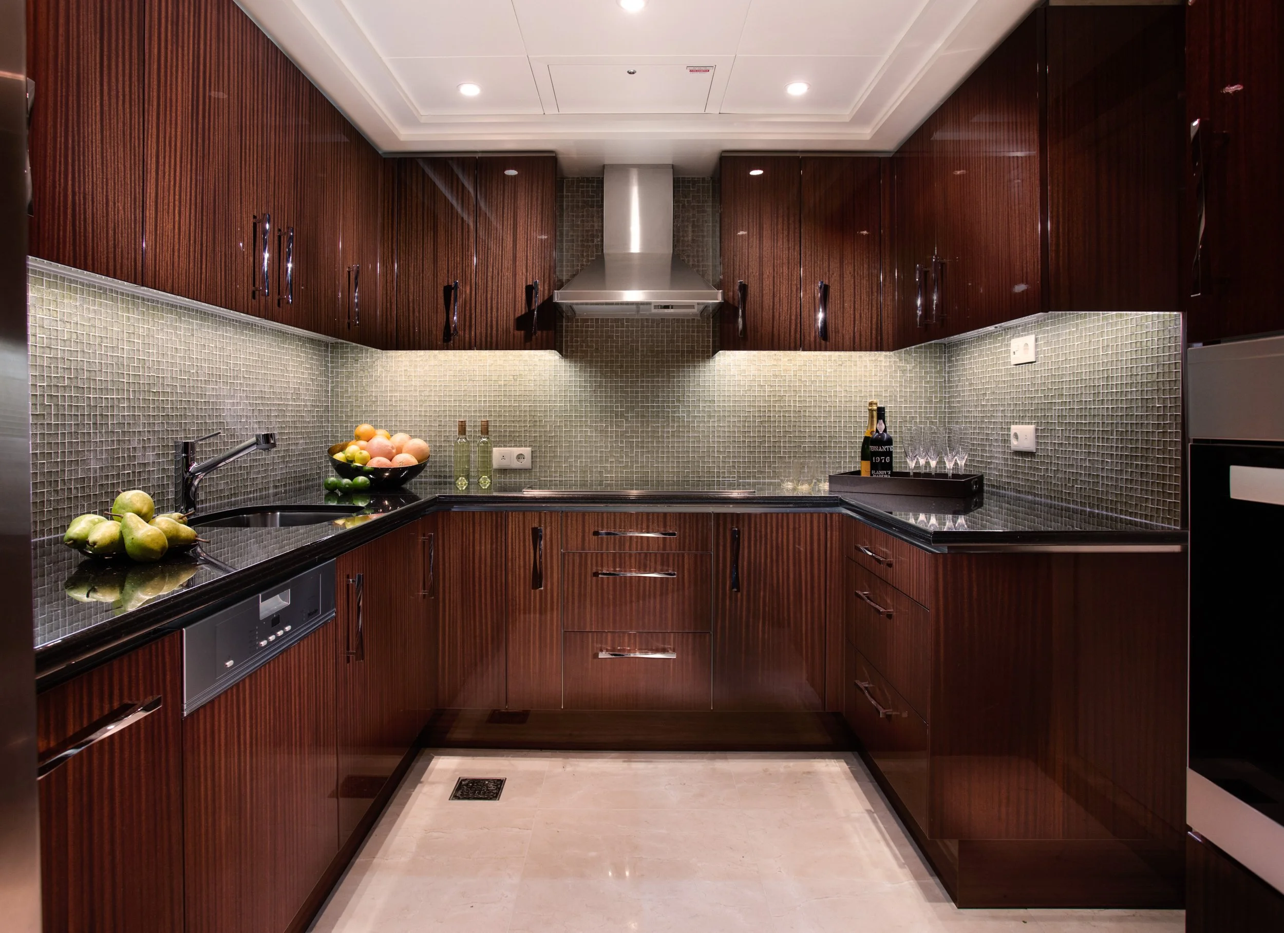 Luxury modern kitchen with dark wood cabinets, gray mosaic tile backsplash, black countertops, stainless steel range hood, fruit bowls, and wine glasses