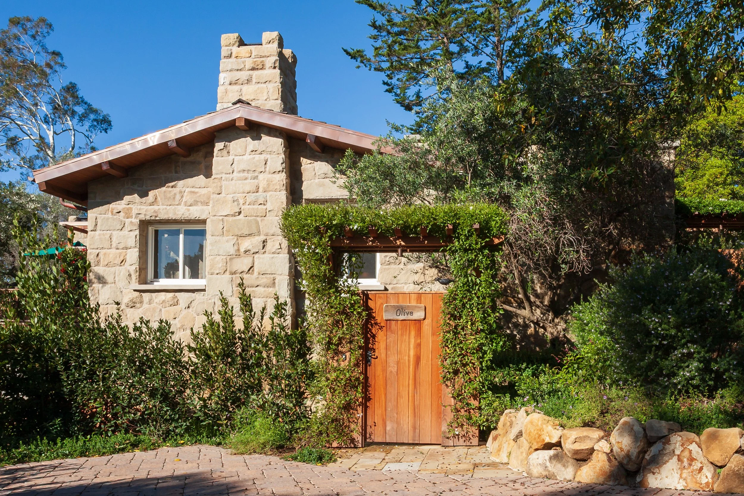 Stone cottage with a chimney, wooden gate labeled Olive, and lush garden landscaping
