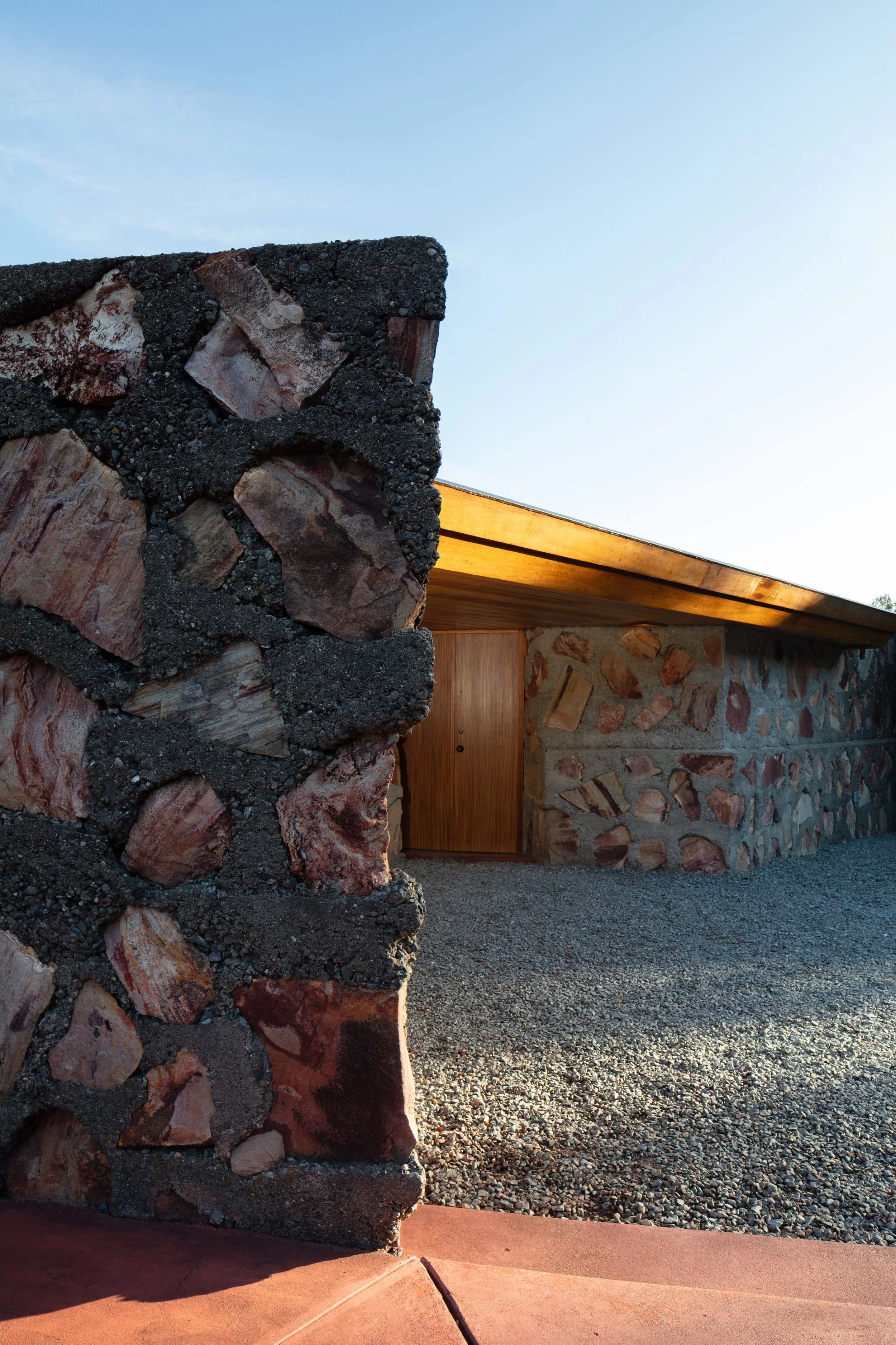 Rustic stone building with a wooden door, dark mortar, and a sloping wood roof under a clear blue sky