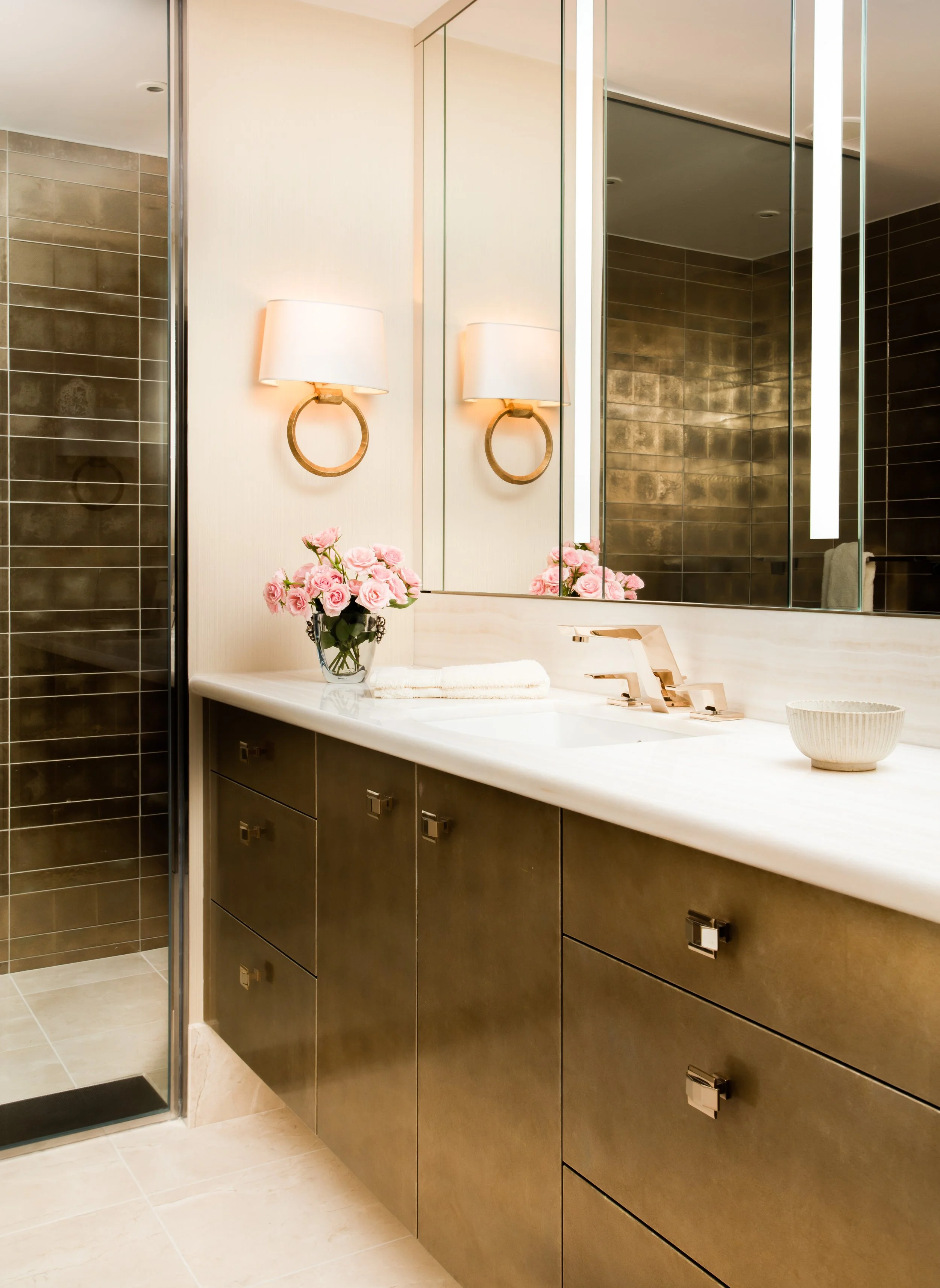 Luxury bathroom vanity with dark tile walls, mirrored cabinets, gold fixtures, white countertop, pink roses, and towels