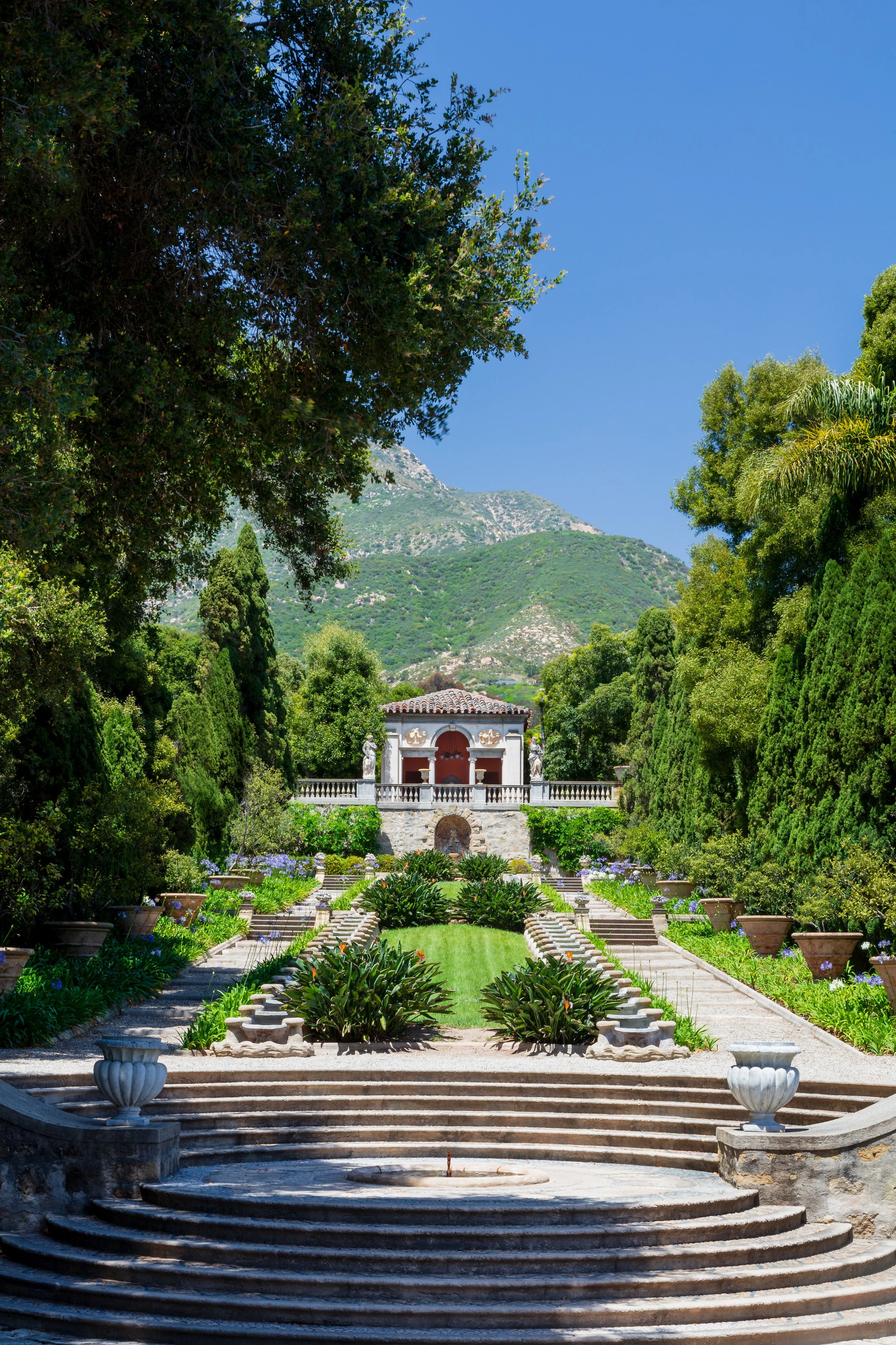 Formal terraced garden with a stone pavilion and fountain, surrounded by lush greenery and mountain views