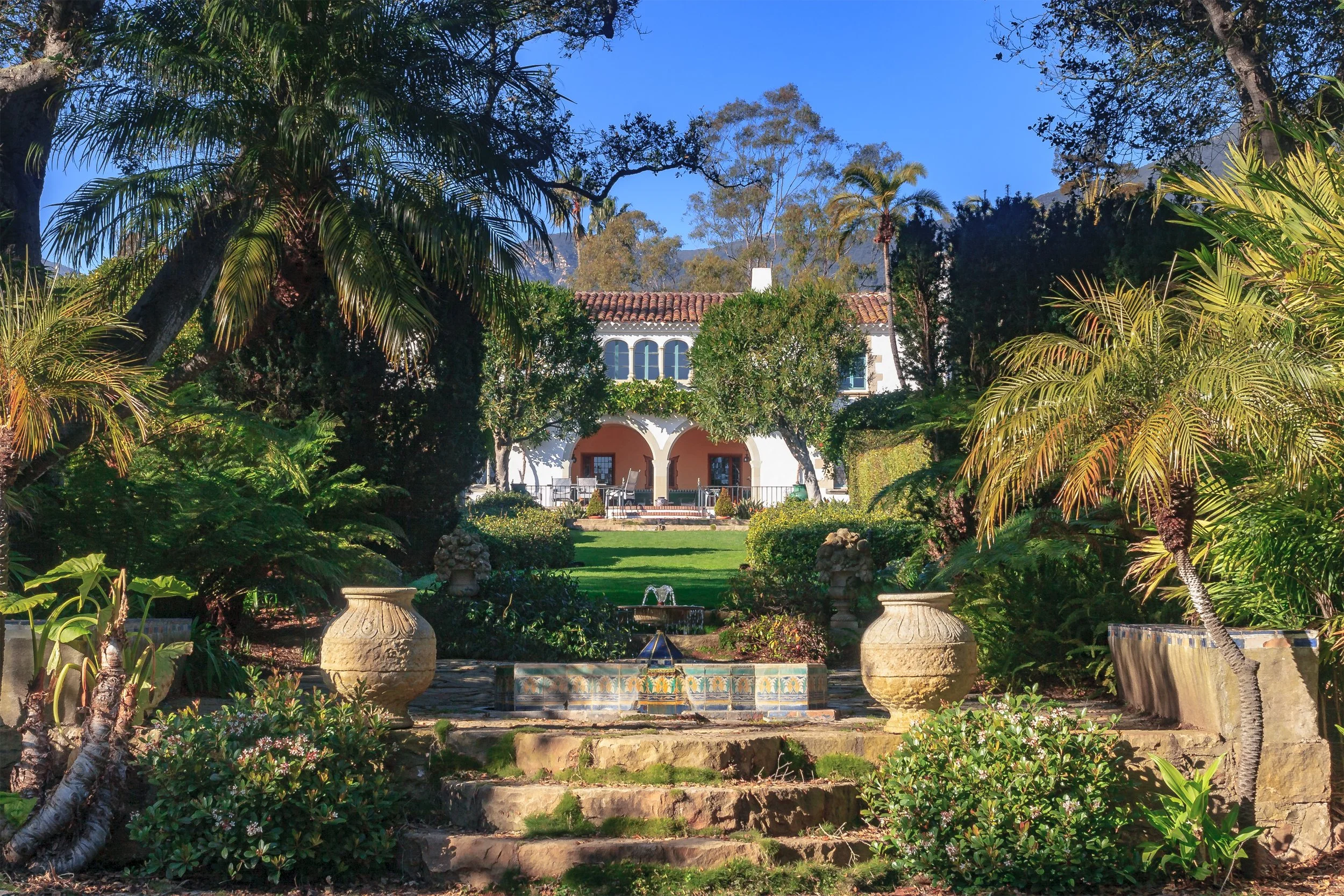 Lush tropical garden with a tiled fountain, palm trees, and Spanish-style villa backed by mountains