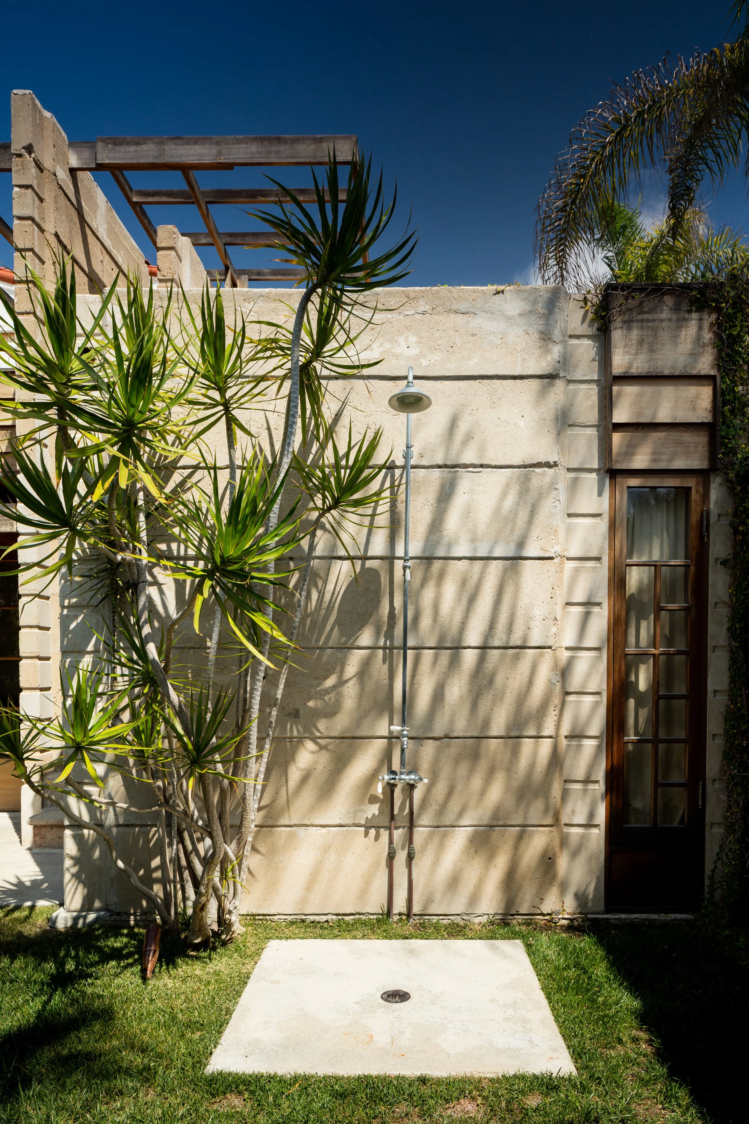 Sunlit courtyard with tropical plants, stucco walls, wooden pergola, outdoor shower, and glass-paneled door