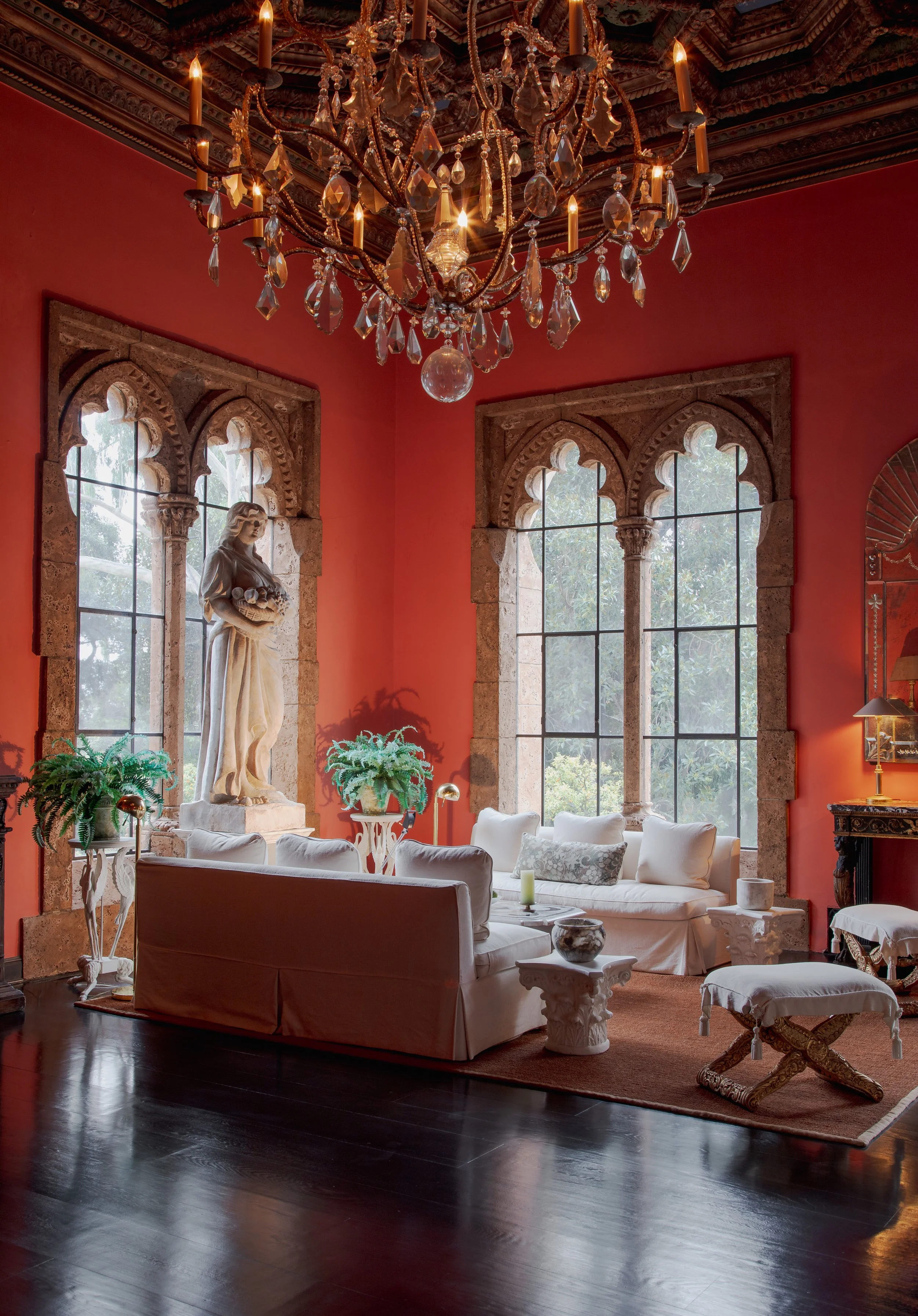 Elegant red-walled living room with a crystal candle chandelier, carved stone windows, classical statue, white sofas, plants, and dark wood floors