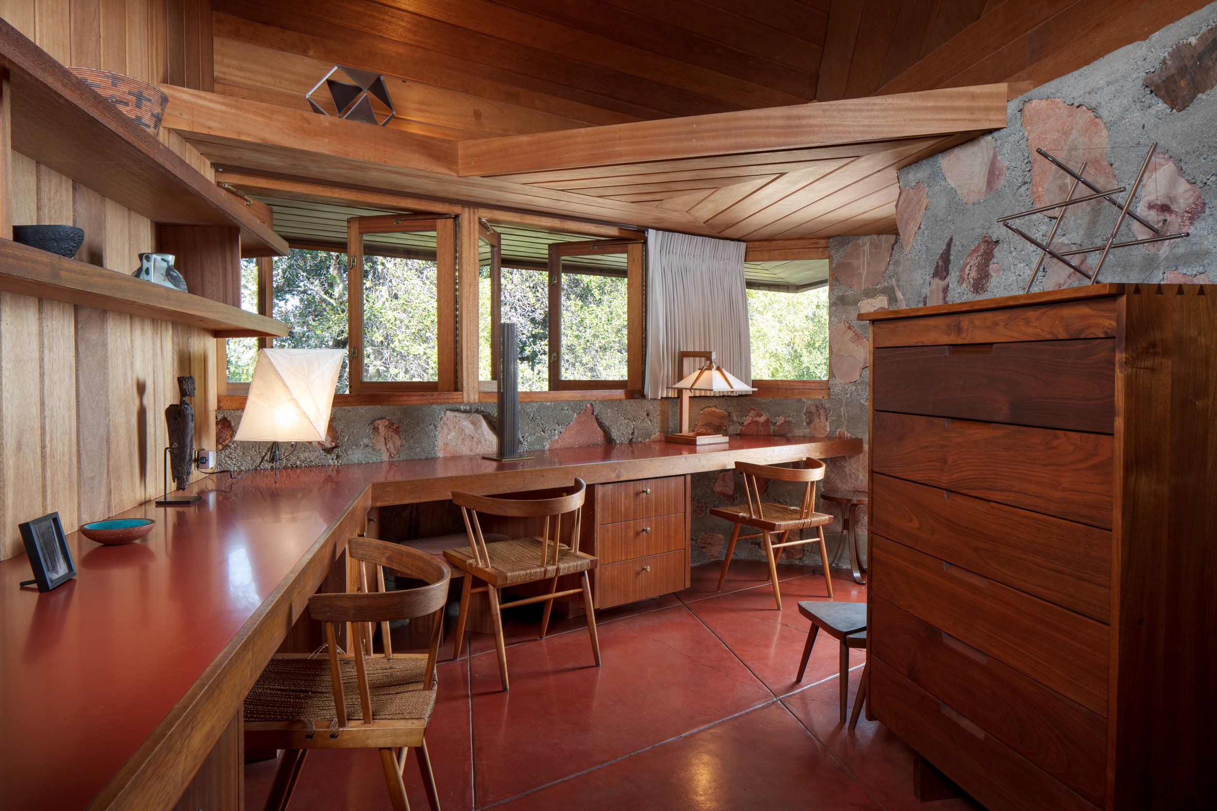 Midcentury modern wood-paneled interior with stone walls, built-in desk, vintage chairs, lamps, and large windows