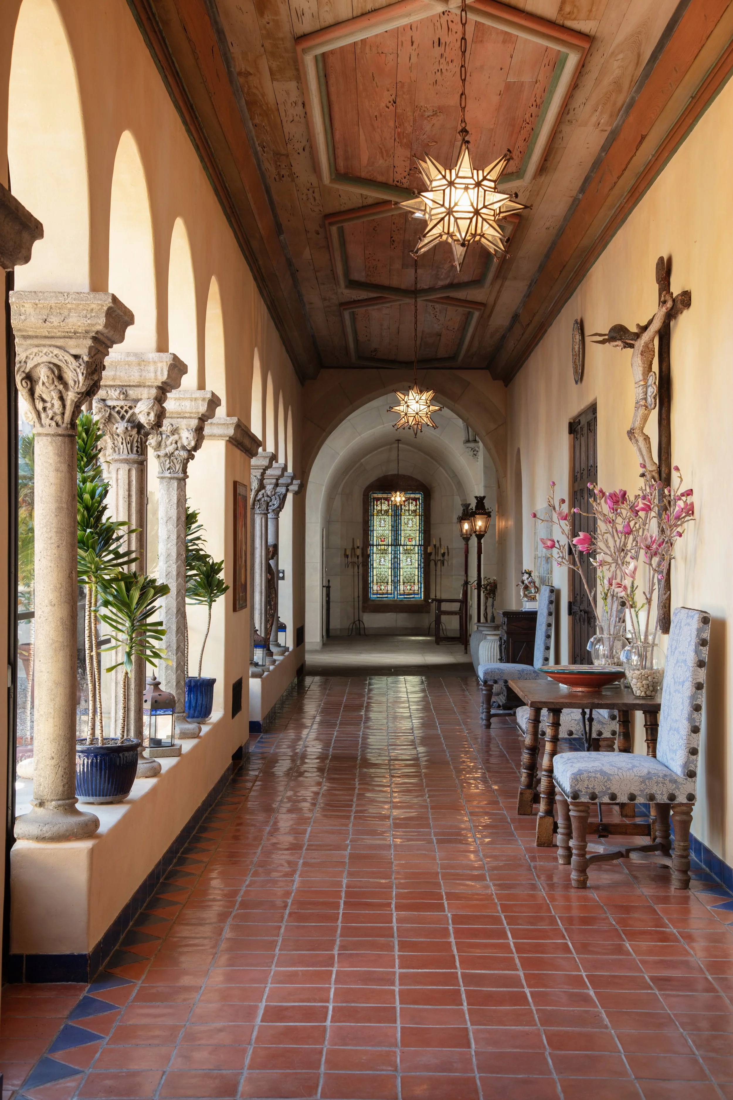 Elegant Spanish-style mansion hallway with terracotta tile floors, carved stone columns, star-shaped pendant lights, stained-glass window, plants, and upholstered chairs