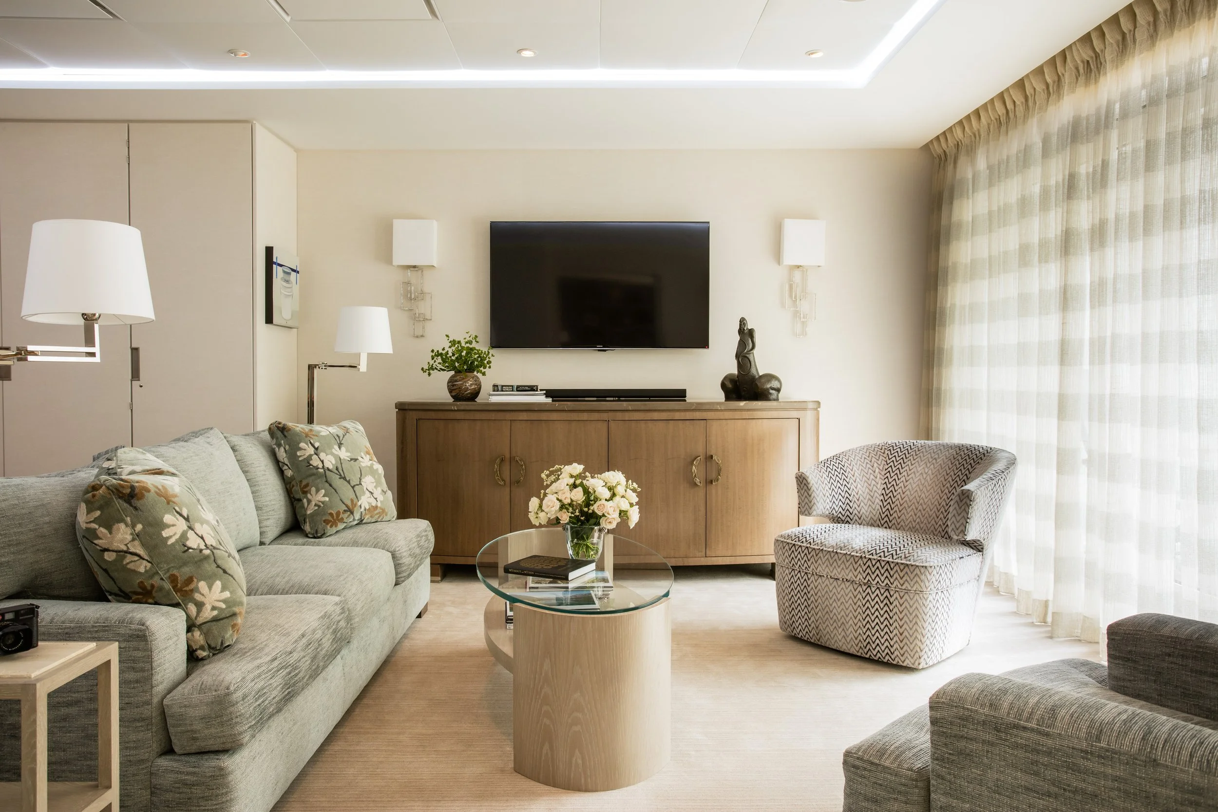 Elegant living room with a wall-mounted TV, wood sideboard, sofas, patterned armchair, glass coffee table, flowers, and sheer curtains.