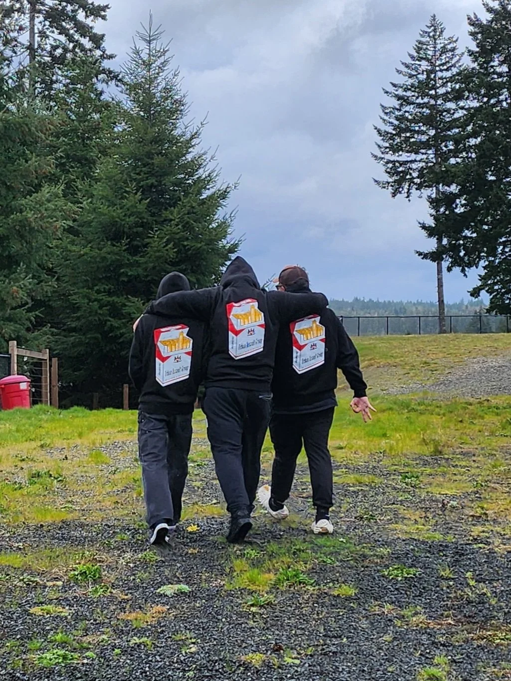 Three people walking outdoors during the daytime, embracing each other, wearing black hoodies with a logo of a white box with yellow and red details on the back.