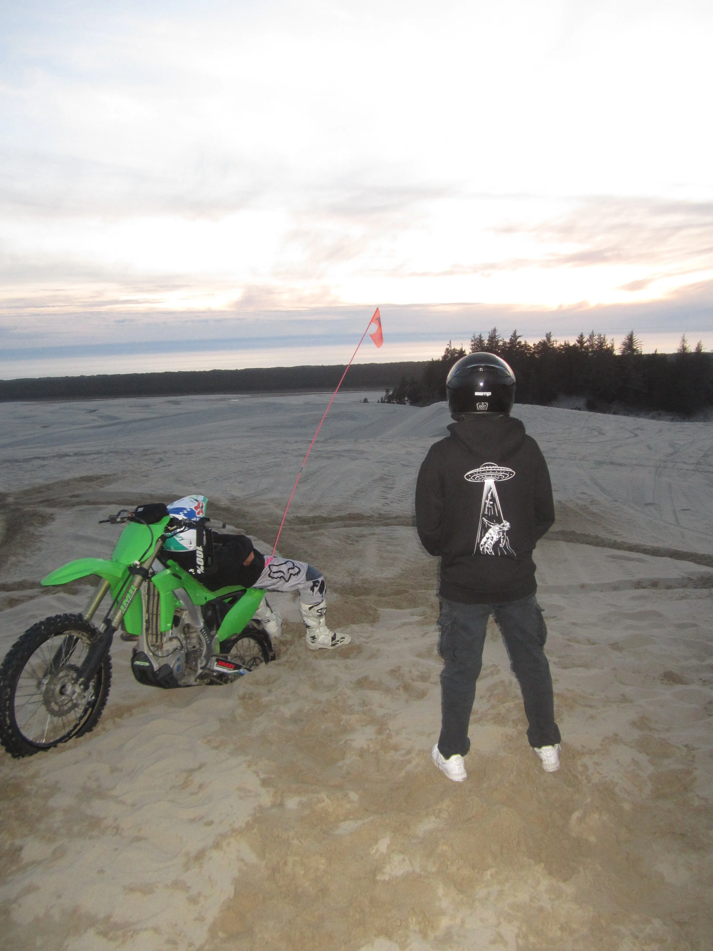 Two people and a dirt bike on a sandy terrain during sunset, with trees and a cloudy sky in the background.