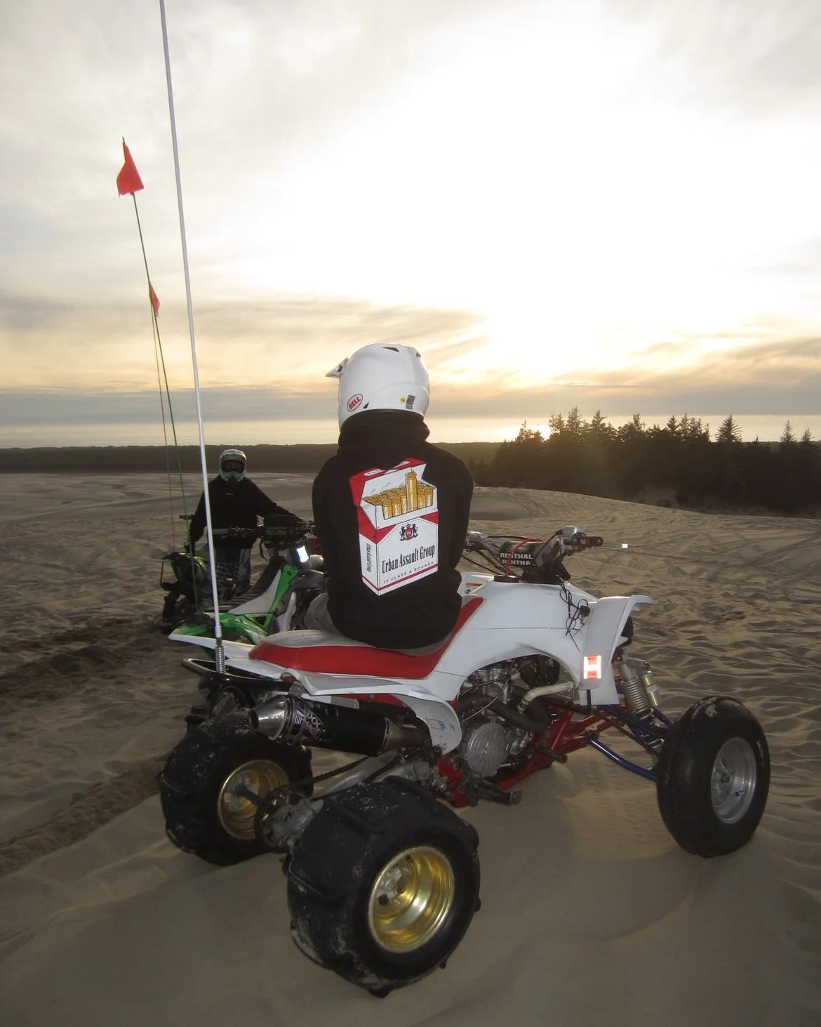 Two off-road motorcycles on a sandy terrain during sunset, with one rider sitting on a white and red ATV and another rider standing nearby. The rider on the ATV wears a black jacket with an 'Urban Assault Group' patch on the back and a white helmet. 