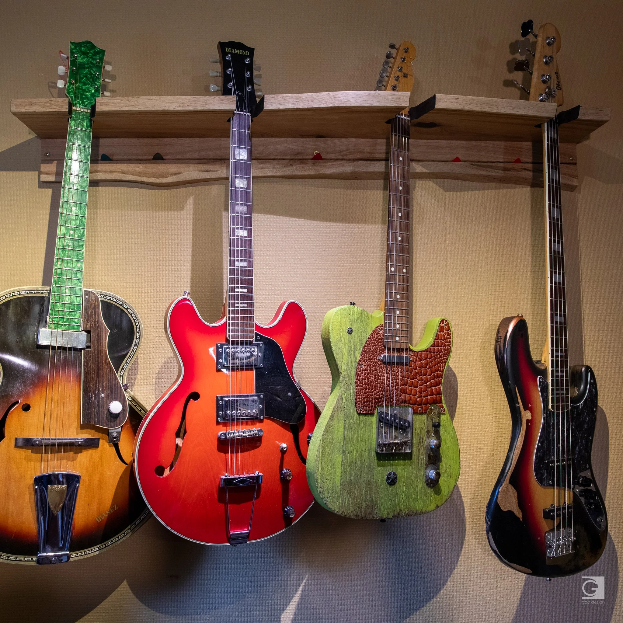 Four electric and acoustic guitars hanging on a wall with a wooden shelf above.