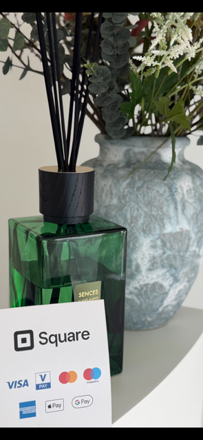 Green square glass diffuser with black reeds, next to a large ceramic vase with white and green plants, and a payment sign displaying various payment options.
