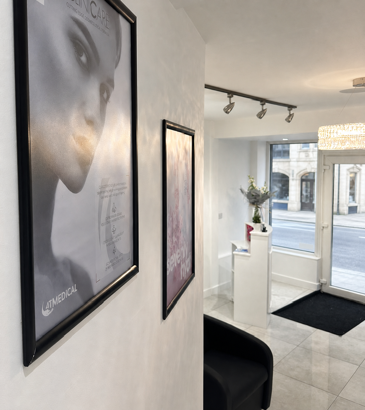Interior of a modern medical or cosmetic clinic waiting area with framed posters on the wall, a black chair, and a white reception desk near a large window and front door, illuminated by a chandelier and ceiling track lights.