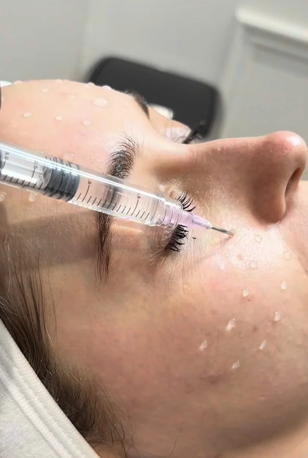 A person receiving a cosmetic procedure, lying down with a syringe near their eye, with water droplets on their face.