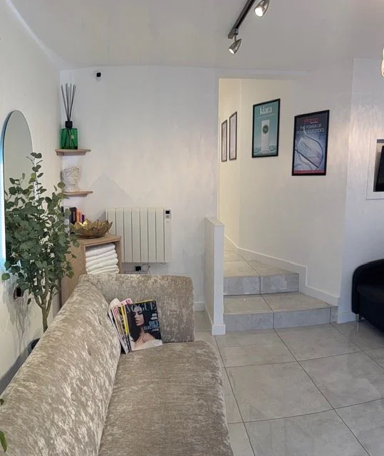 A cozy interior living room with a beige plush sofa holding magazines, a green potted plant, a wall mirror, and small wall-mounted shelves with decorative items, leading to a short staircase and framed posters on the wall.