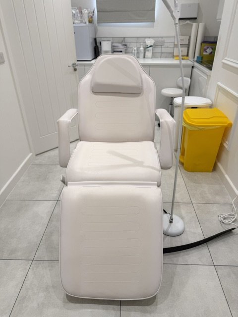 Empty white medical examination chair in a clinical room with gray tiled floor, medical supplies on a counter, a yellow waste bin, and medical equipment.
