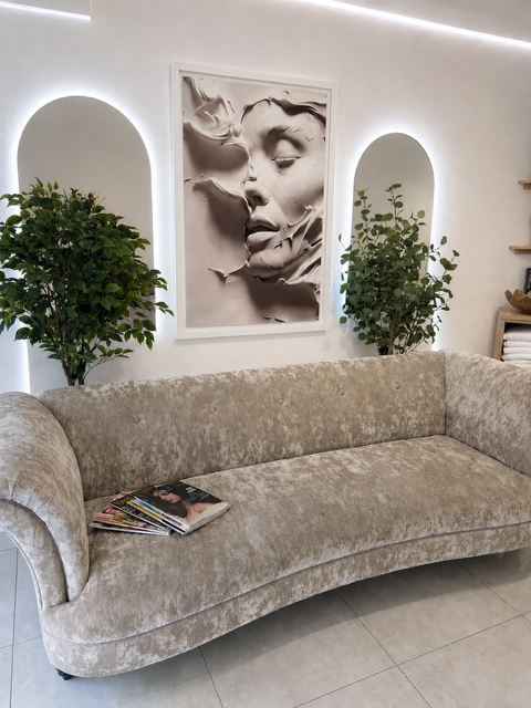 A beige velvet sofa with a stack of magazines on one end in a modern interior with white walls, two potted plants, and a large black-and-white artistic portrait of a woman's face on the wall.