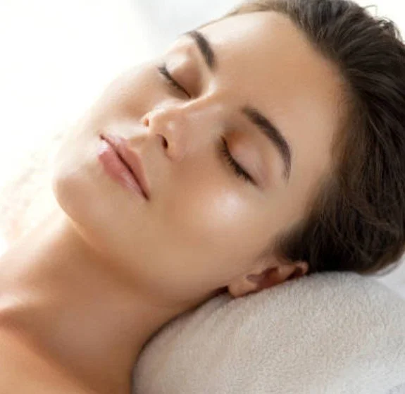 Close-up of a woman with closed eyes, relaxed facial expression, lying down on a pillow with her head turned slightly to the side after a facial.