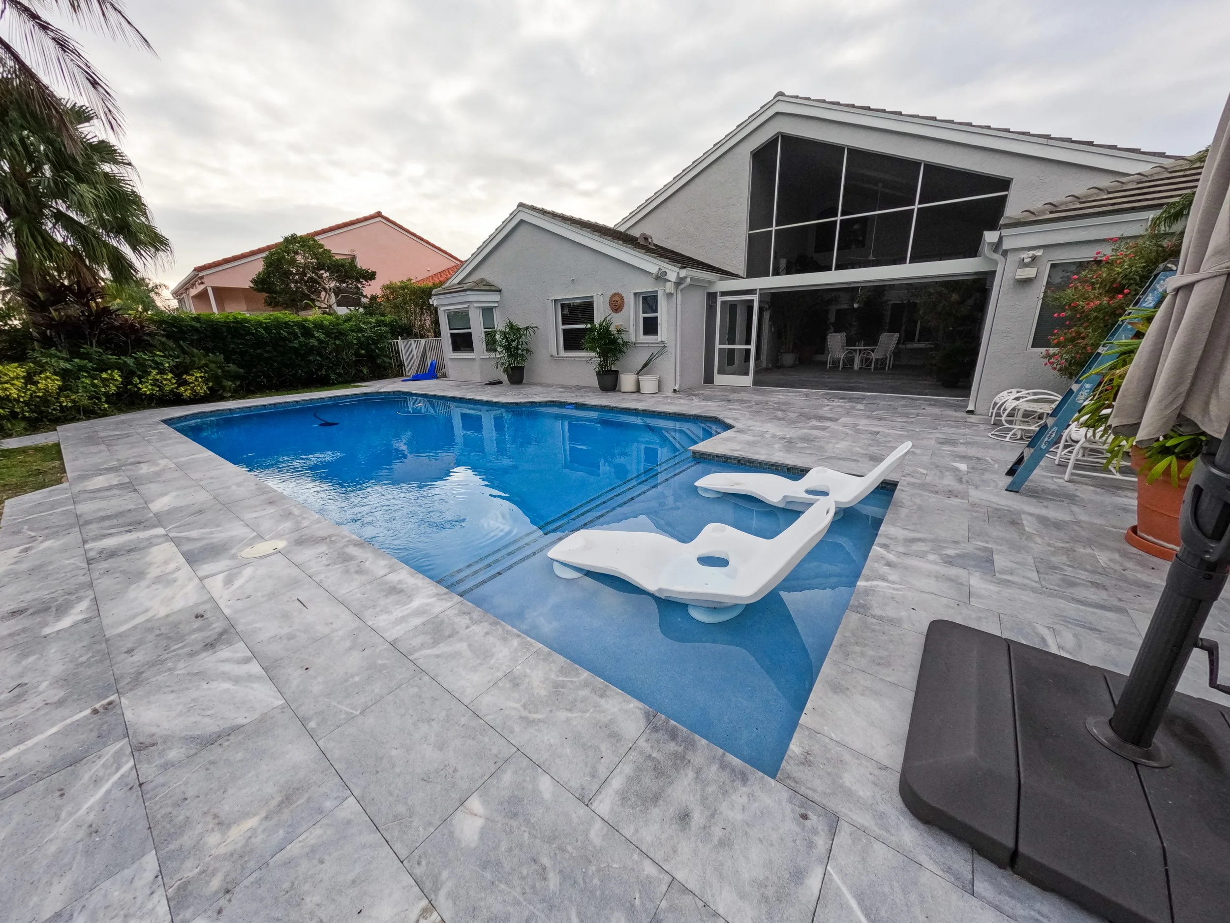 Backyard with a gray stone patio and a swimming pool, featuring two white lounge chairs in the shallow end. The area is surrounded by a white house with a screened porch, lush greenery, and various outdoor furniture.