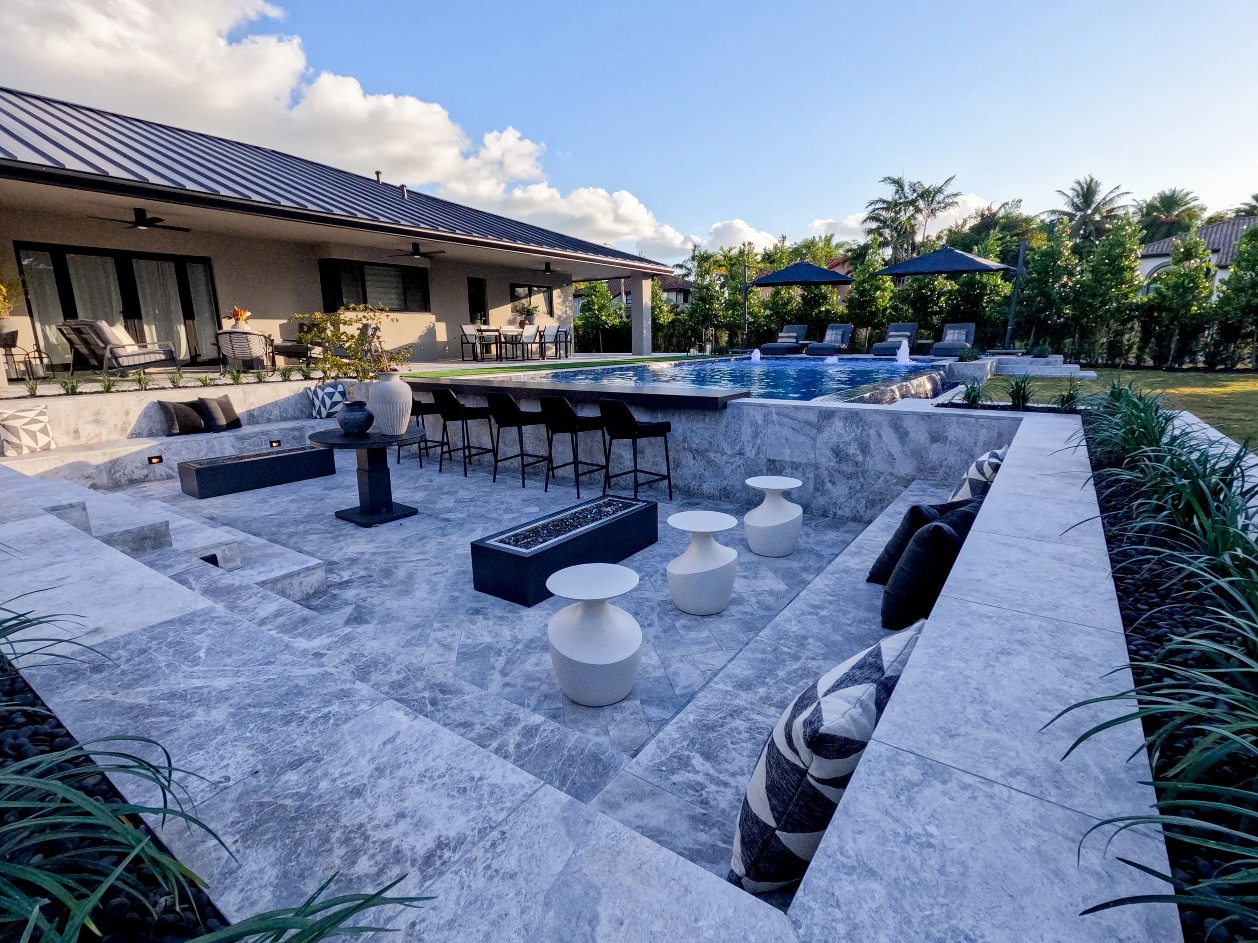 Luxury backyard with swimming pool, poolside chairs, umbrellas, outdoor dining area, and modern house with solar panels under a blue sky with clouds.  built in avenir, fl by the top pool company hammond products