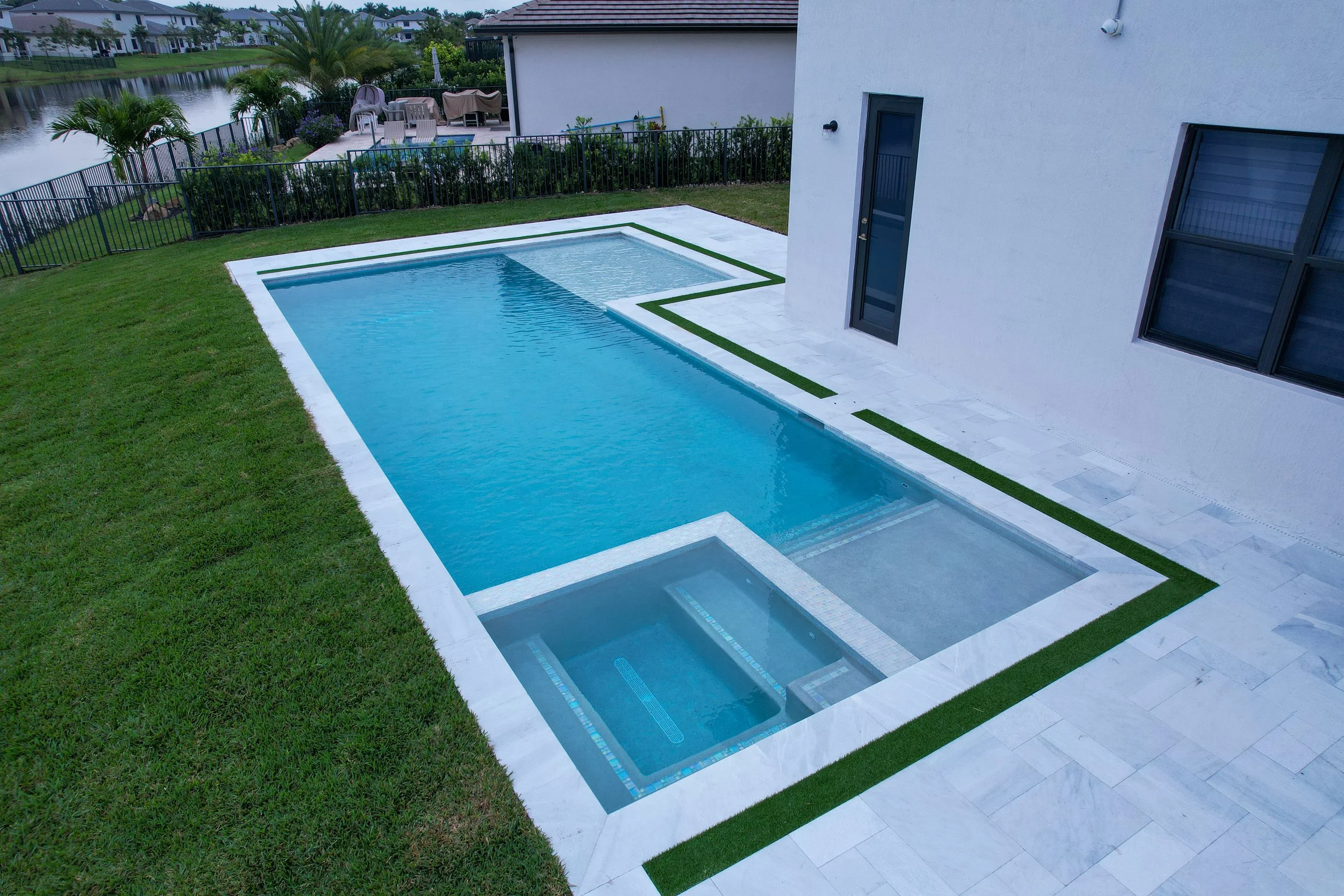 Modern backyard with rectangular swimming pool and hot tub, surrounded by a tiled deck and grass, adjacent to a white house.