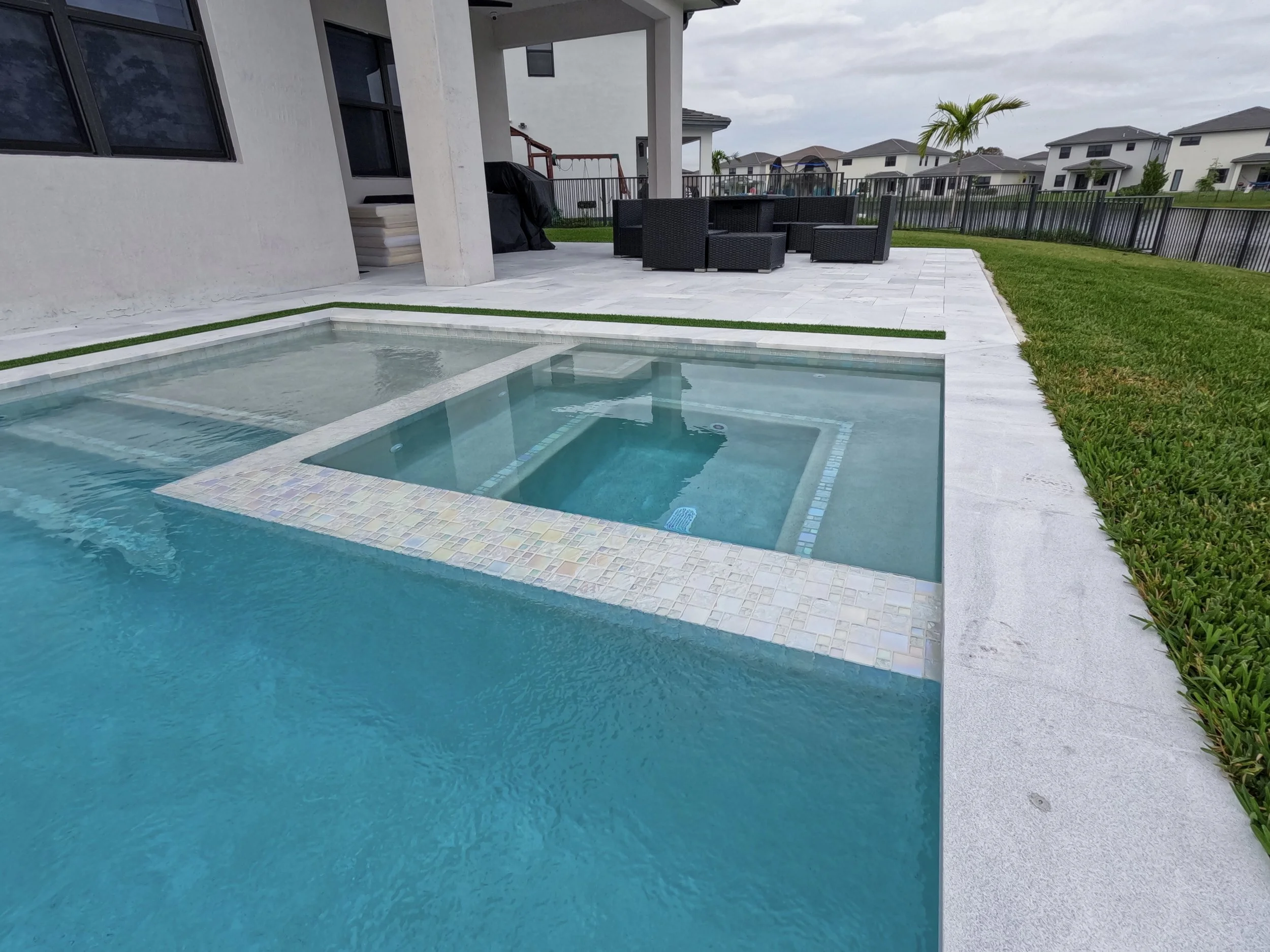 Modern backyard with a swimming pool, spa, and patio furniture. The pool has a separate elevated spa section. The patio area includes wicker furniture and is surrounded by a lawn and fence, with houses visible in the background.