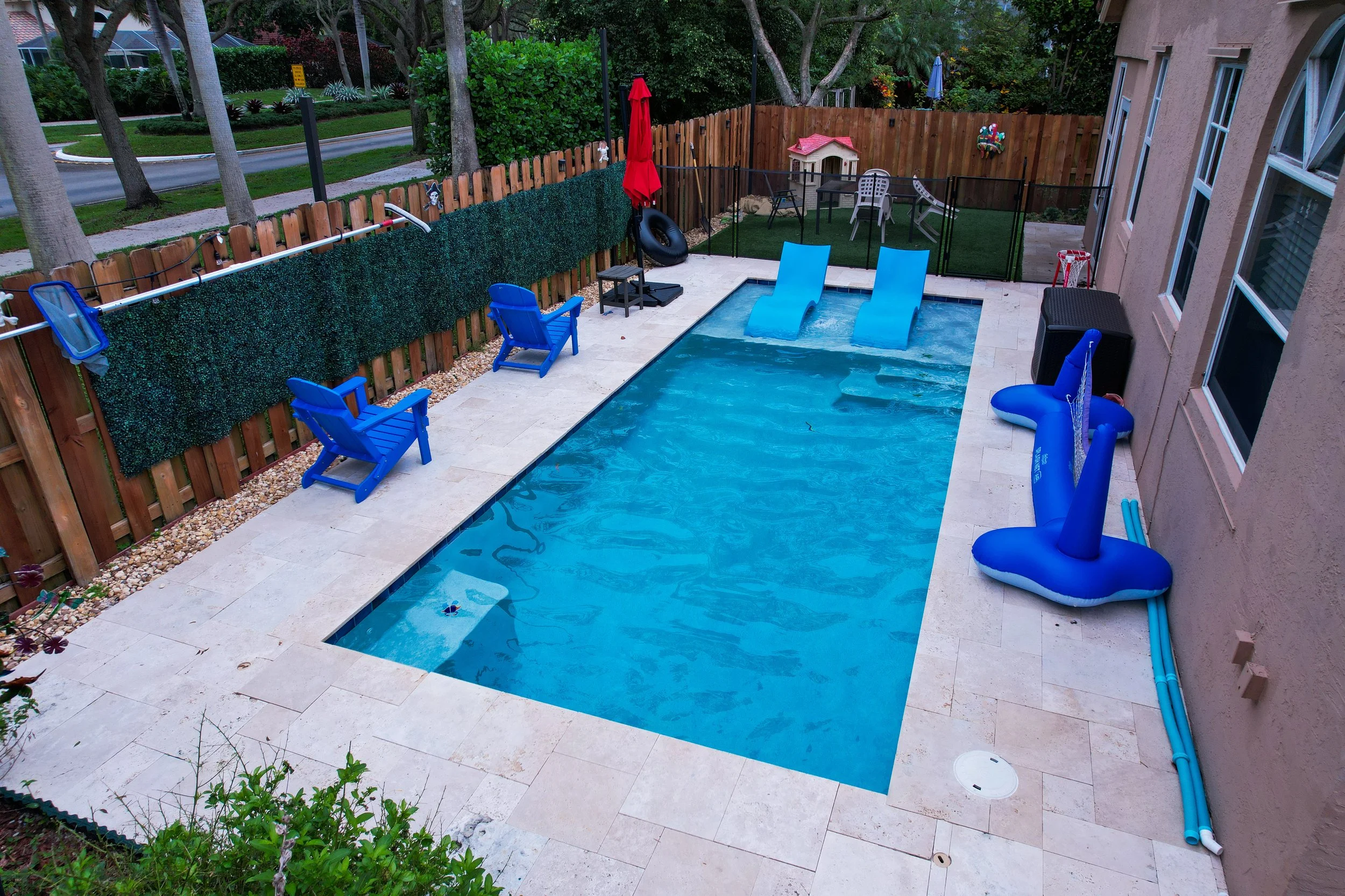 Residential backyard swimming pool with blue lounge chairs, inflatable pool toys, two blue chairs, and a small playhouse on the surrounding patio area.